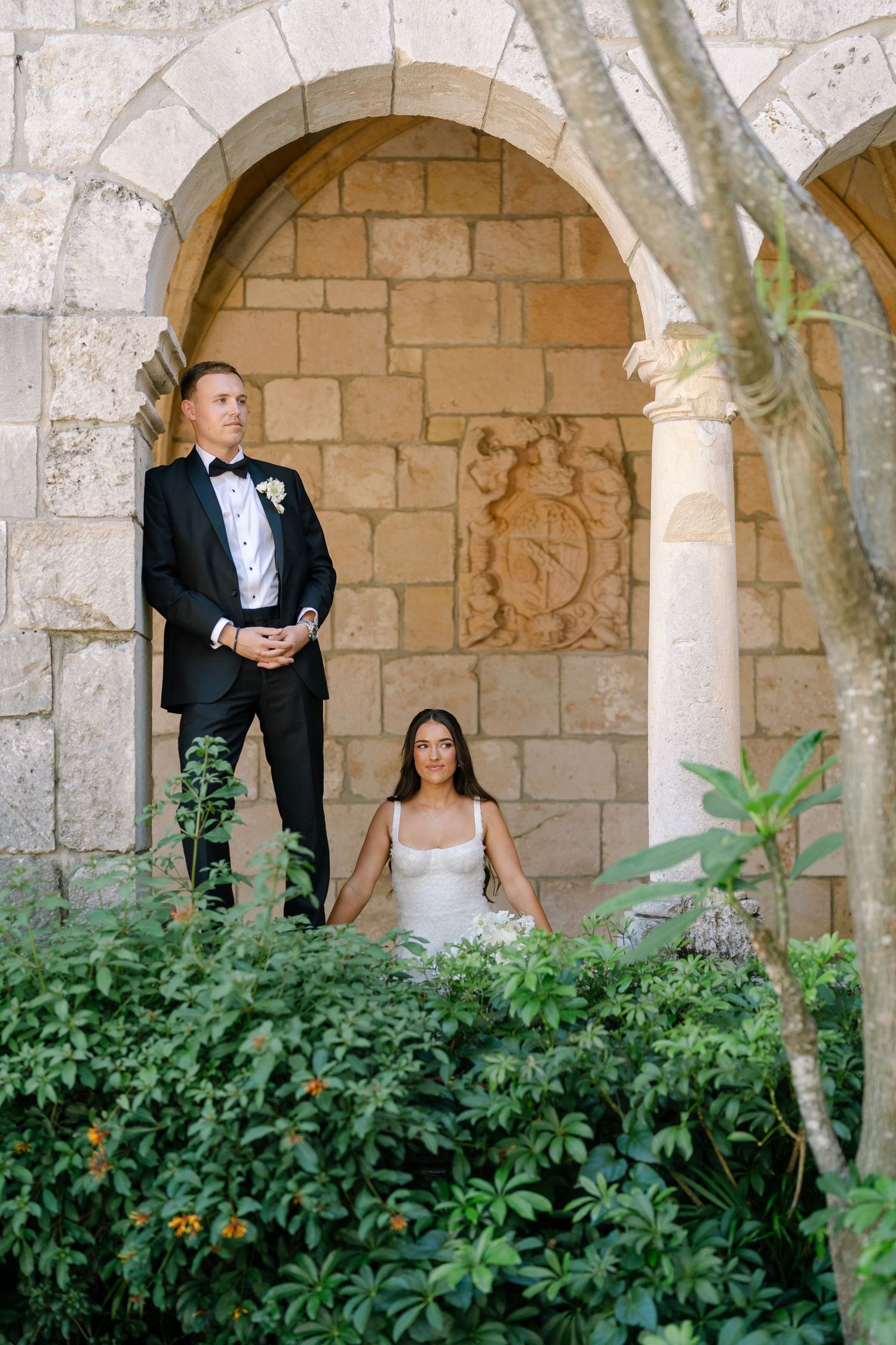 A bride in white and groom in black tuxedo pose together in a stone archway surrounded by lush greenery.