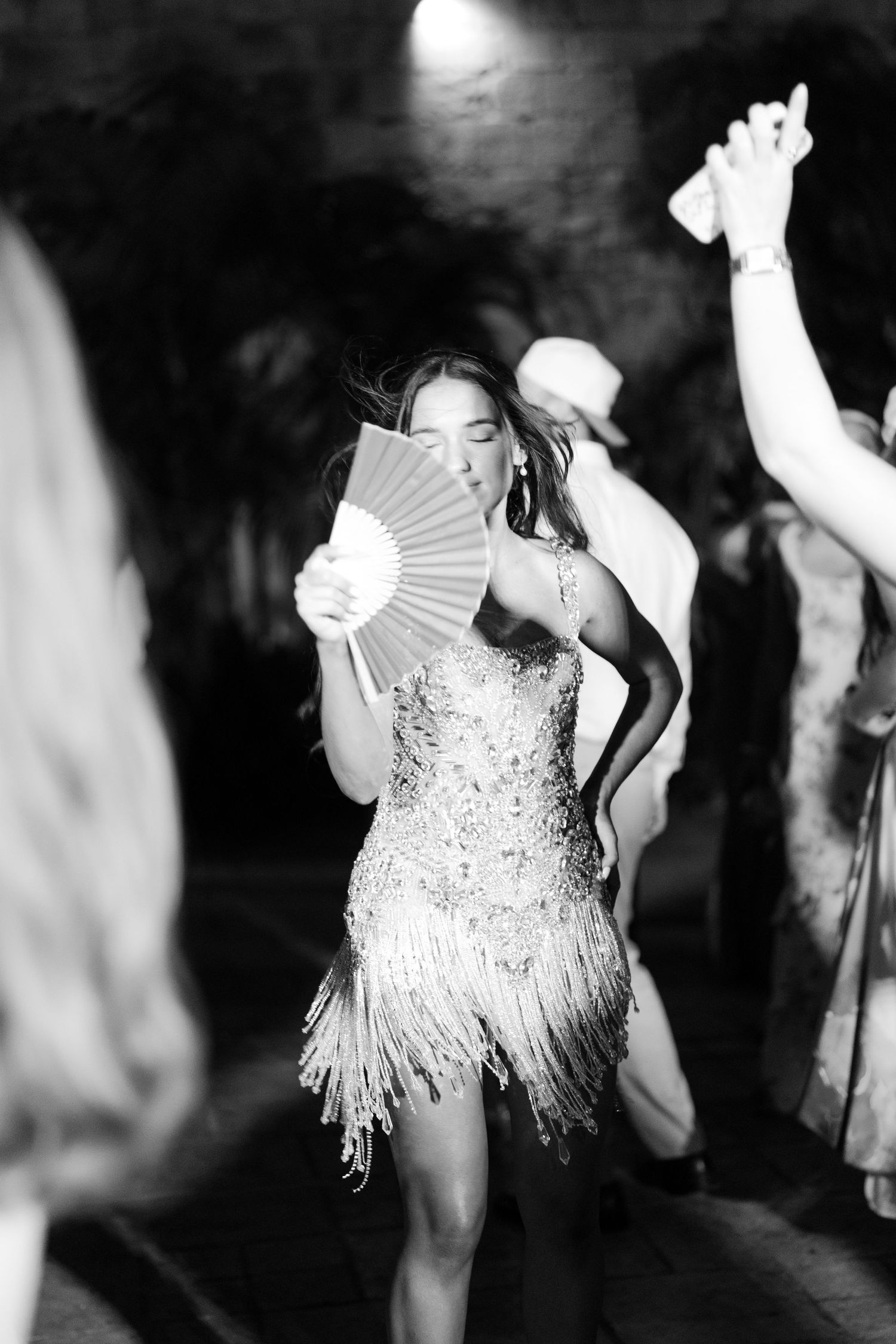 Bride dancing in sparkly fringe reception dress holding white fan at wedding party, black and white.
