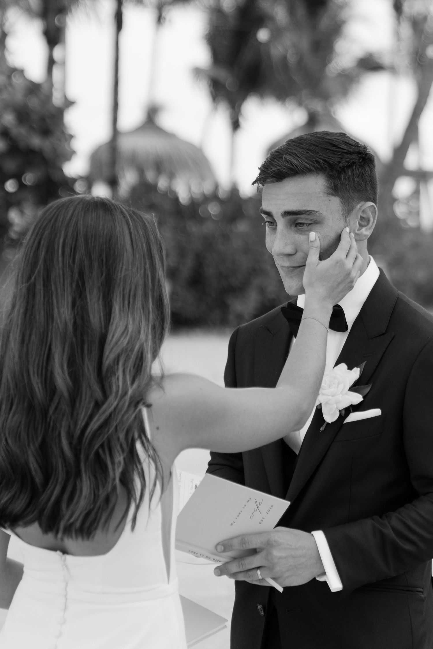 Groom emotional as bride wipes tear from his cheek during outdoor wedding ceremony with palm trees.