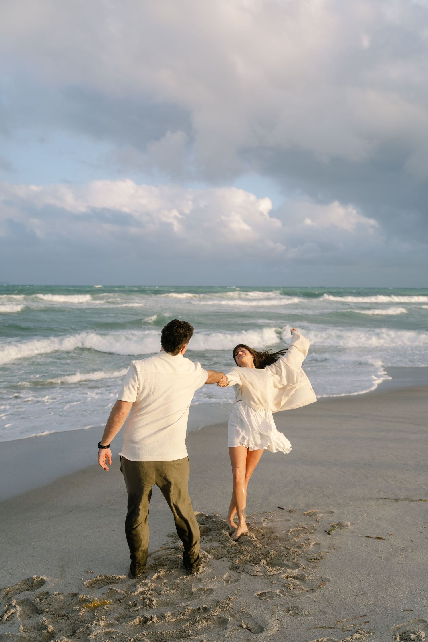 A couple walks along a stormy beach holding hands and leaning into the strong ocean wind together.