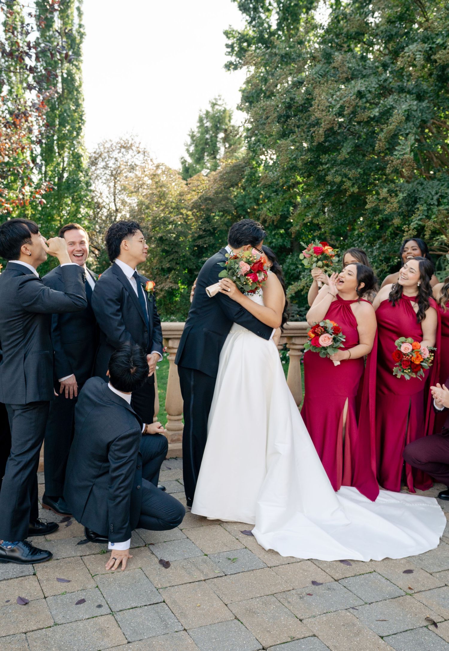 Wedding party in red dresses and black suits poses playfully on stone terrace.