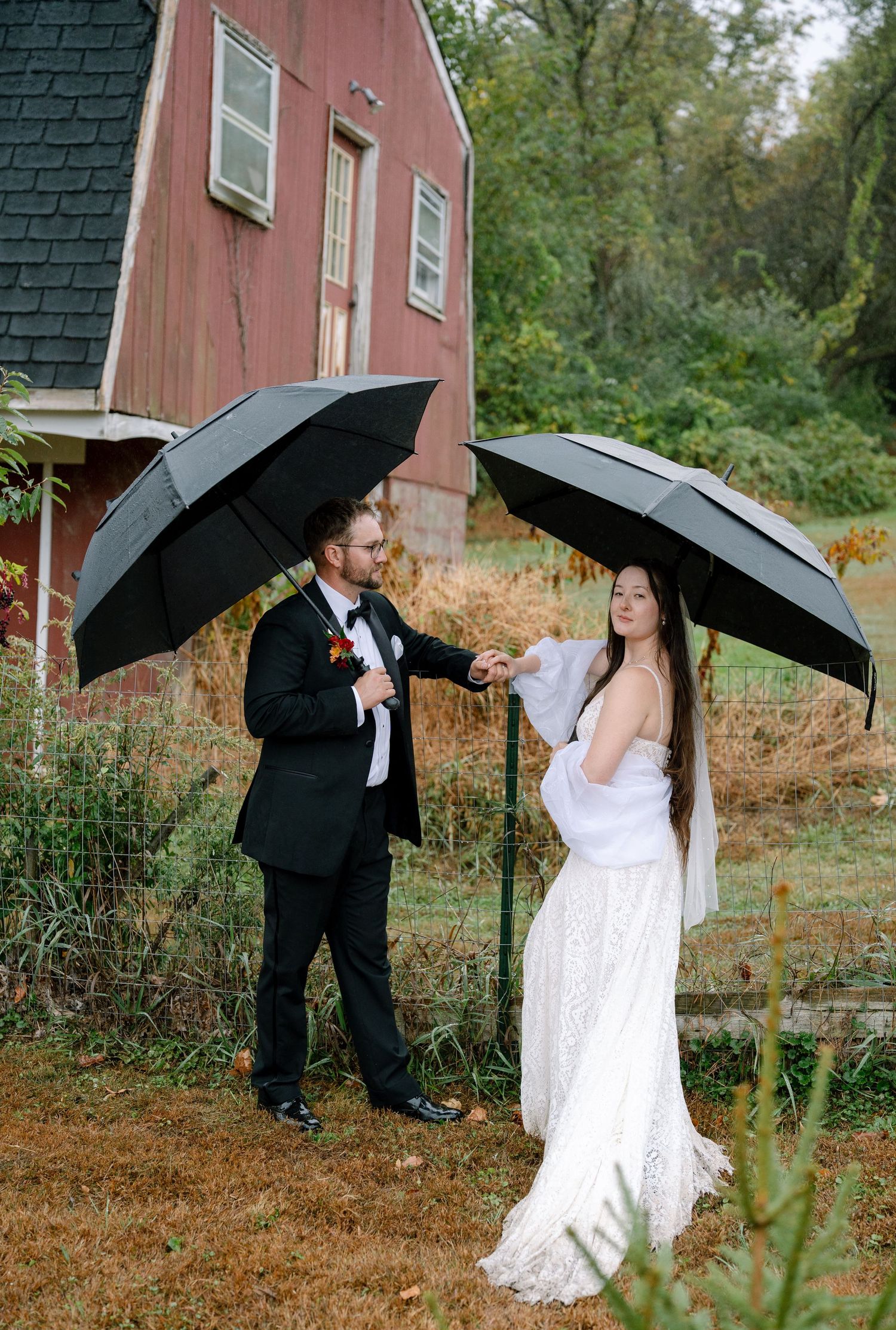 A couple with black umbrellas poses in front of a red barn on their rainy wedding day.