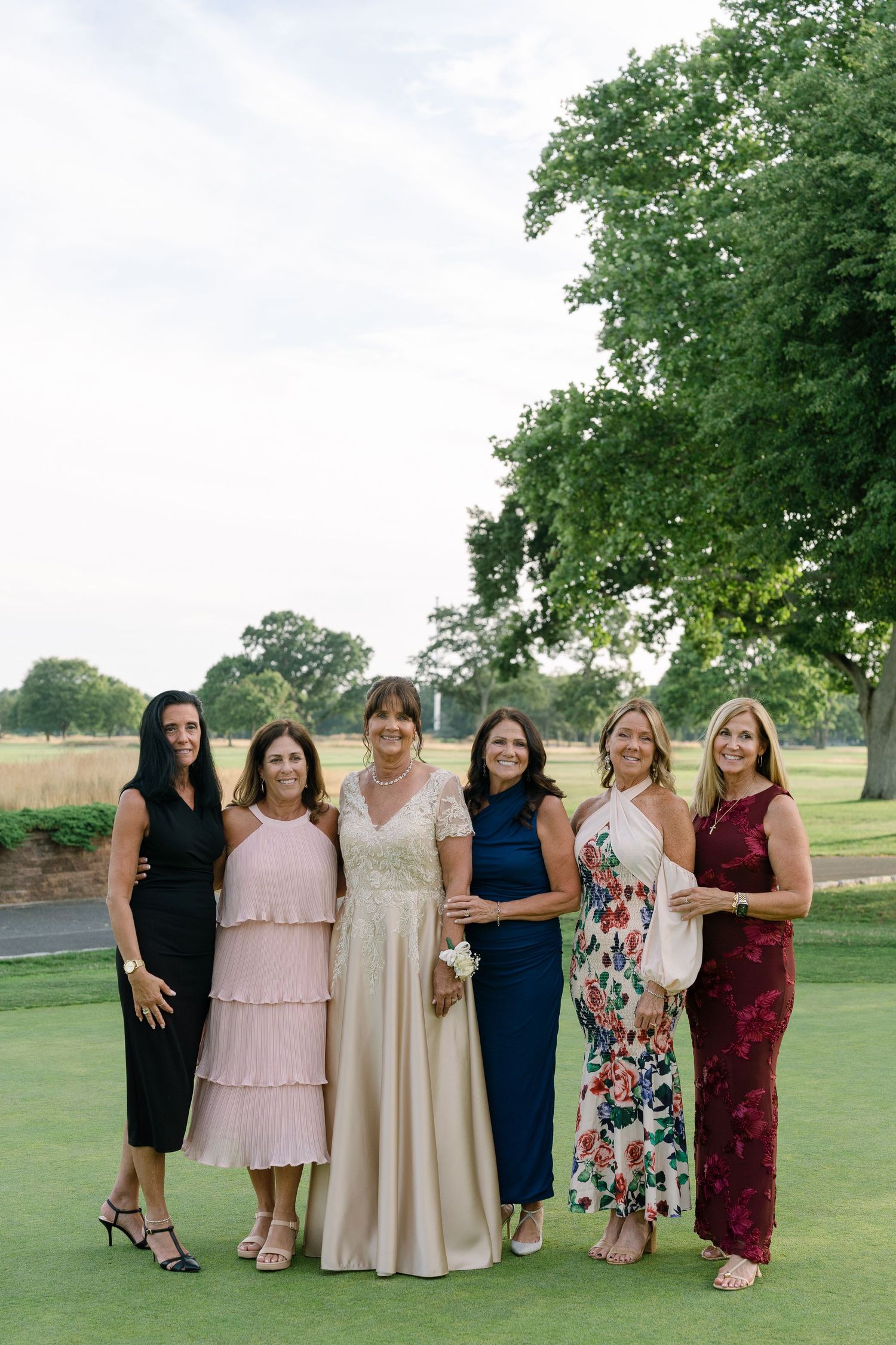 Six women in elegant dresses pose together on a golf course at a wedding reception.