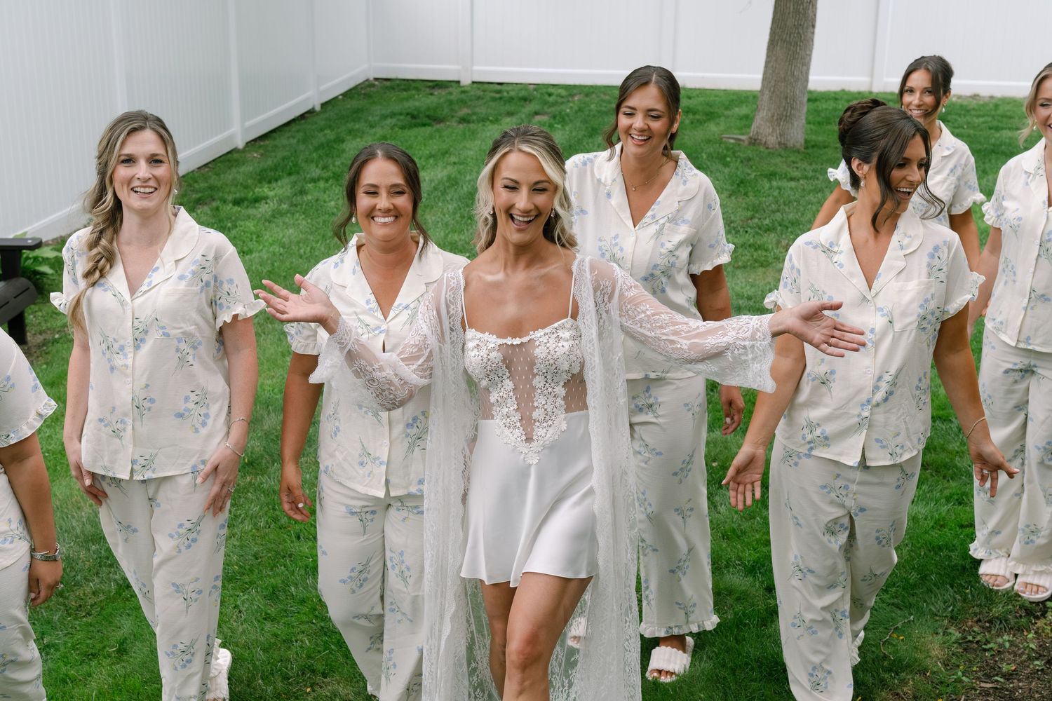 Bride in white mini dress walks with bridesmaids wearing matching floral pajamas on green lawn.