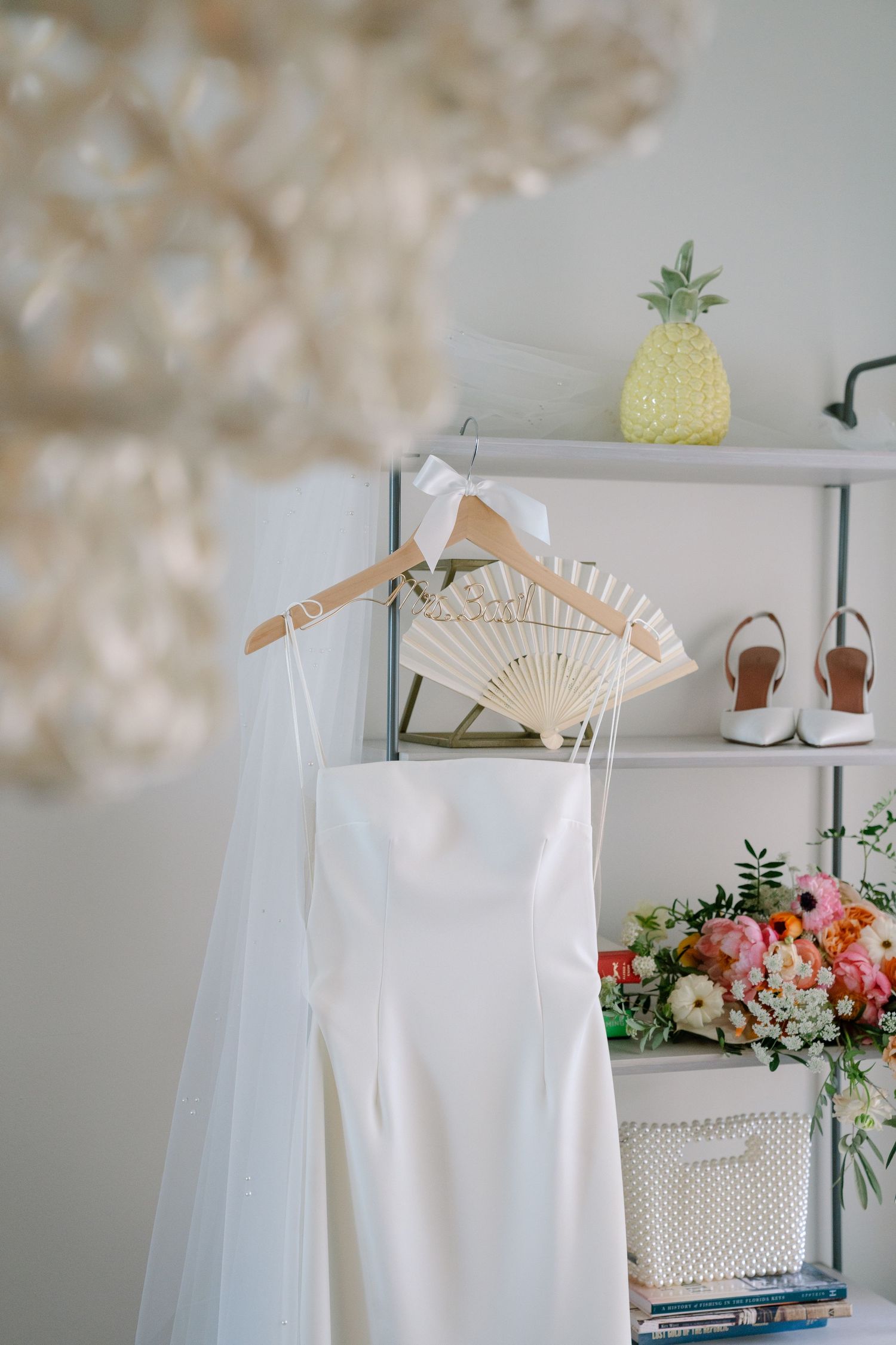 White wedding dress hanging on wooden hanger with floral arrangements and pineapple decor in bright interior setting.