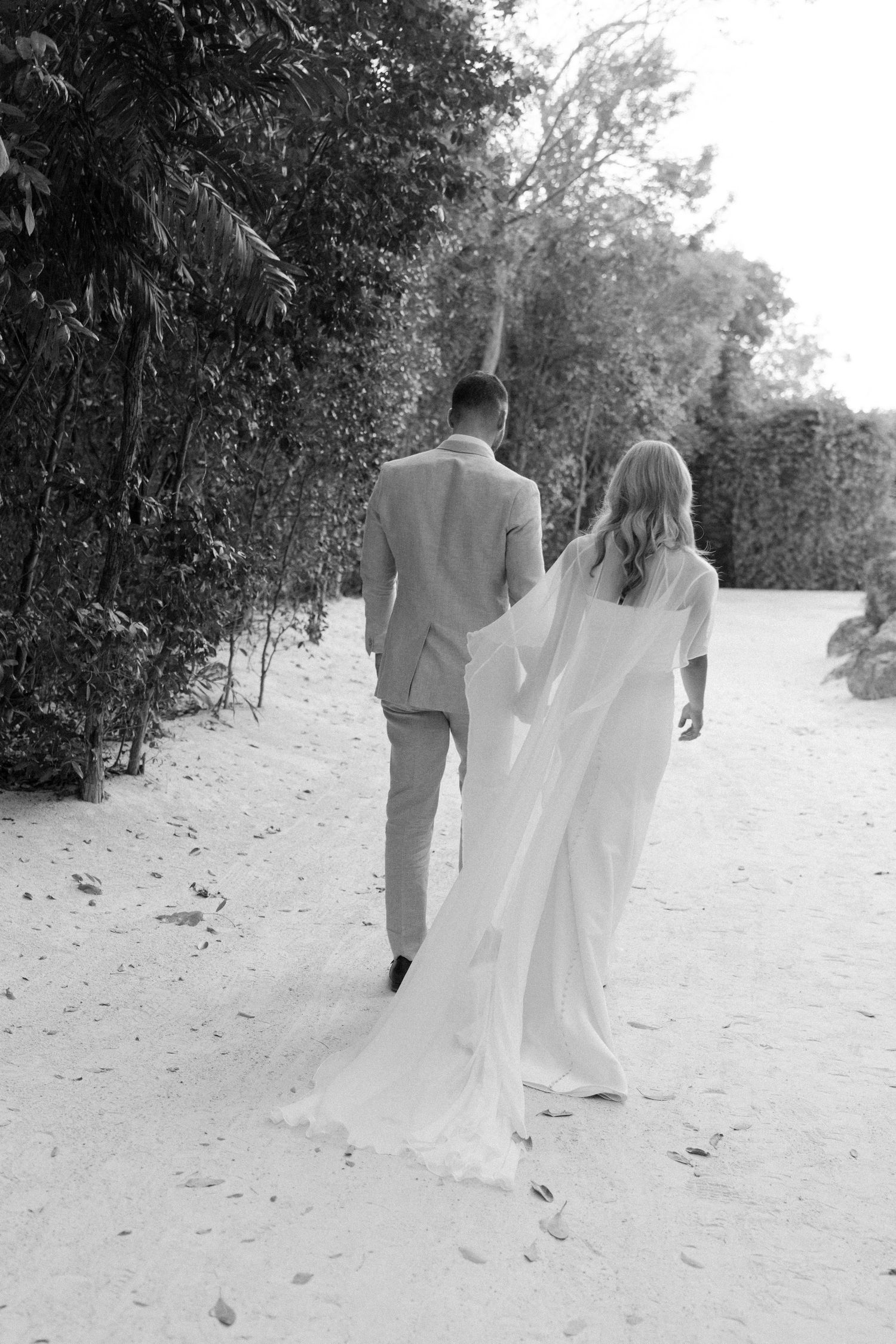 Black and white photo of newlyweds walking along a beach path bordered by tropical foliage.