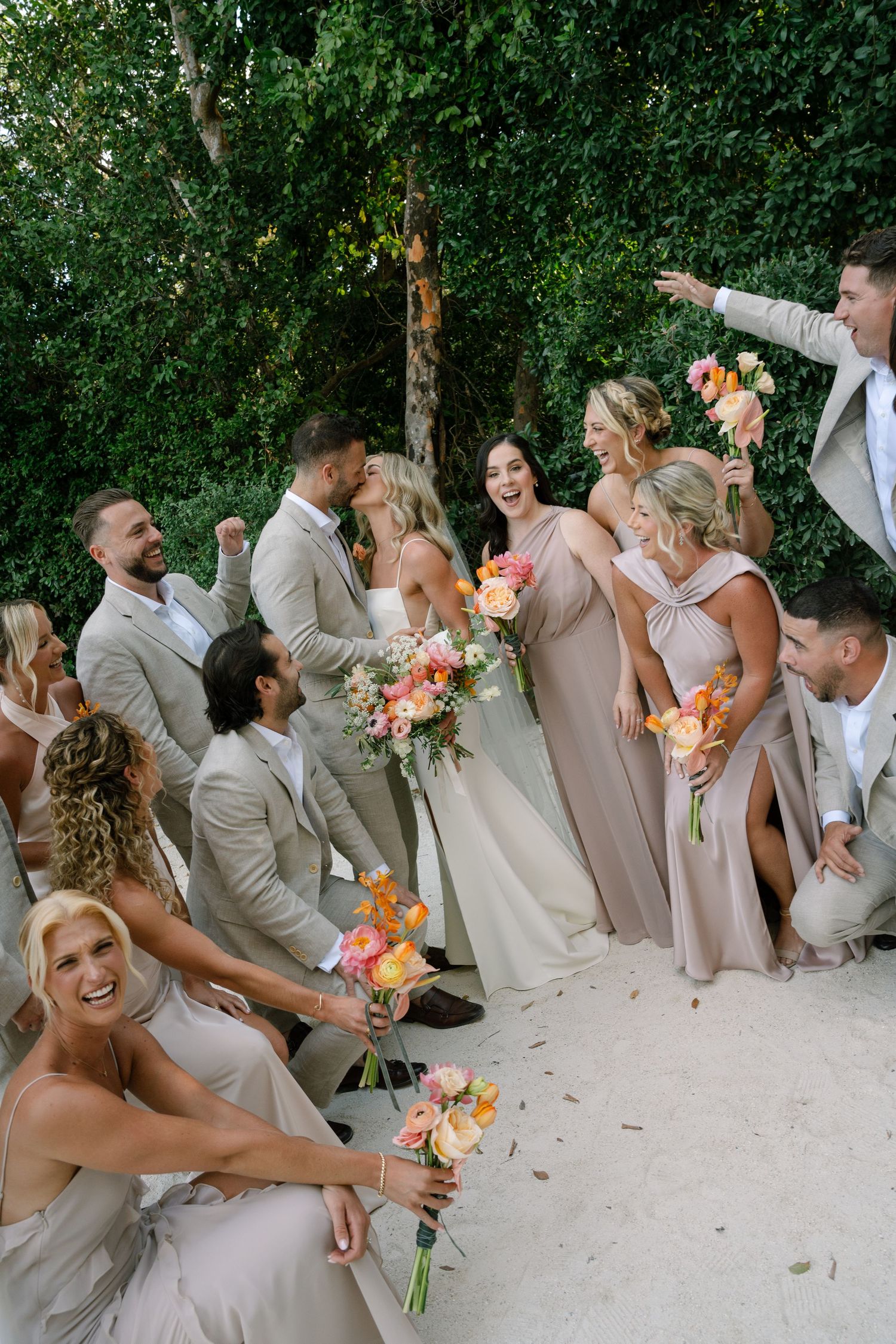 Wedding party in beige and pink attire poses playfully with bouquets against lush greenery backdrop.