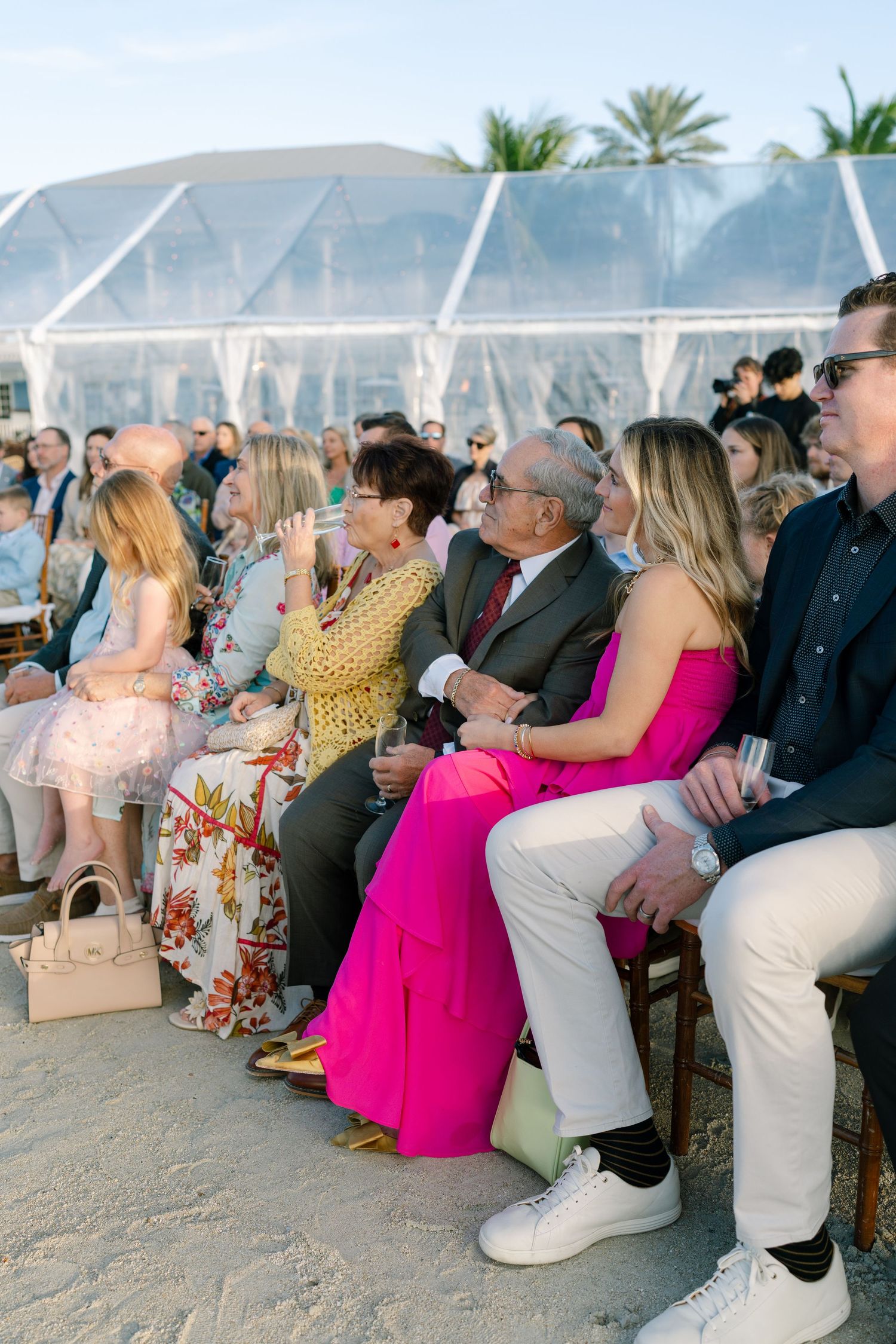 Wedding ceremony guests seated in white chairs on a beach with clear tent visible.