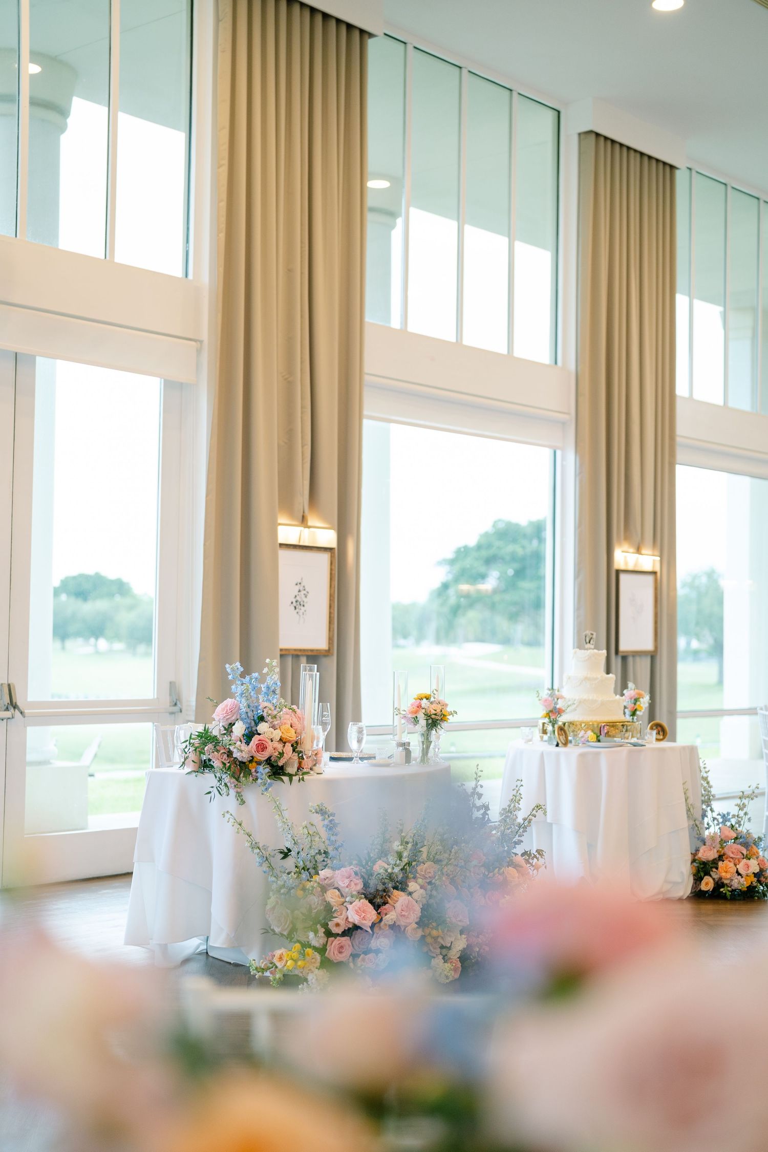 Elegant reception venue with tall windows, beige curtains, and pastel floral centerpieces on white tablecloths.
