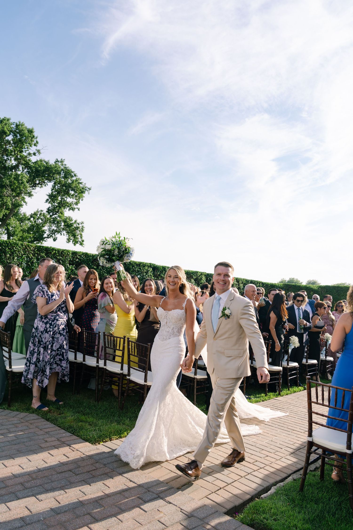 Joyful bride and groom walk back down the aisle after outdoor wedding ceremony with guests cheering.