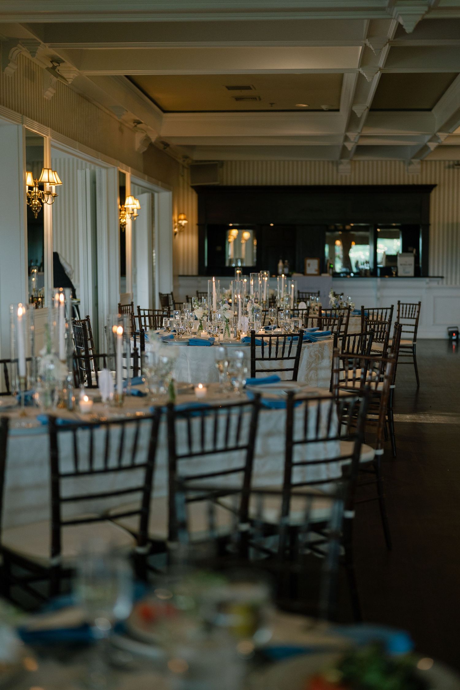 Elegant wedding reception hall with dark Chiavari chairs, candlelit centerpieces, and blue accents on white-draped tables.