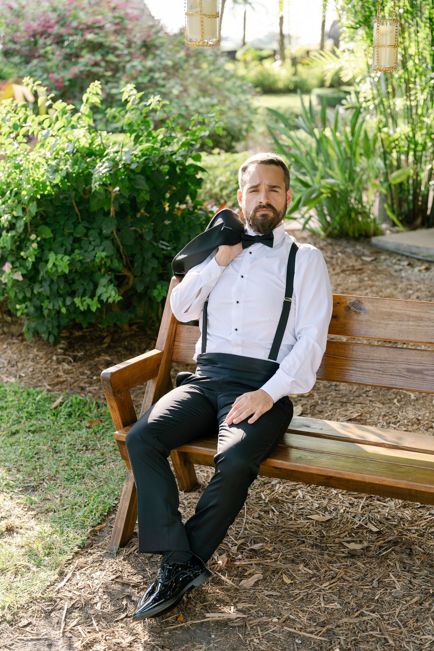A man with a beard wearing a white shirt and suspenders sits on a wooden bench in a lush garden.
