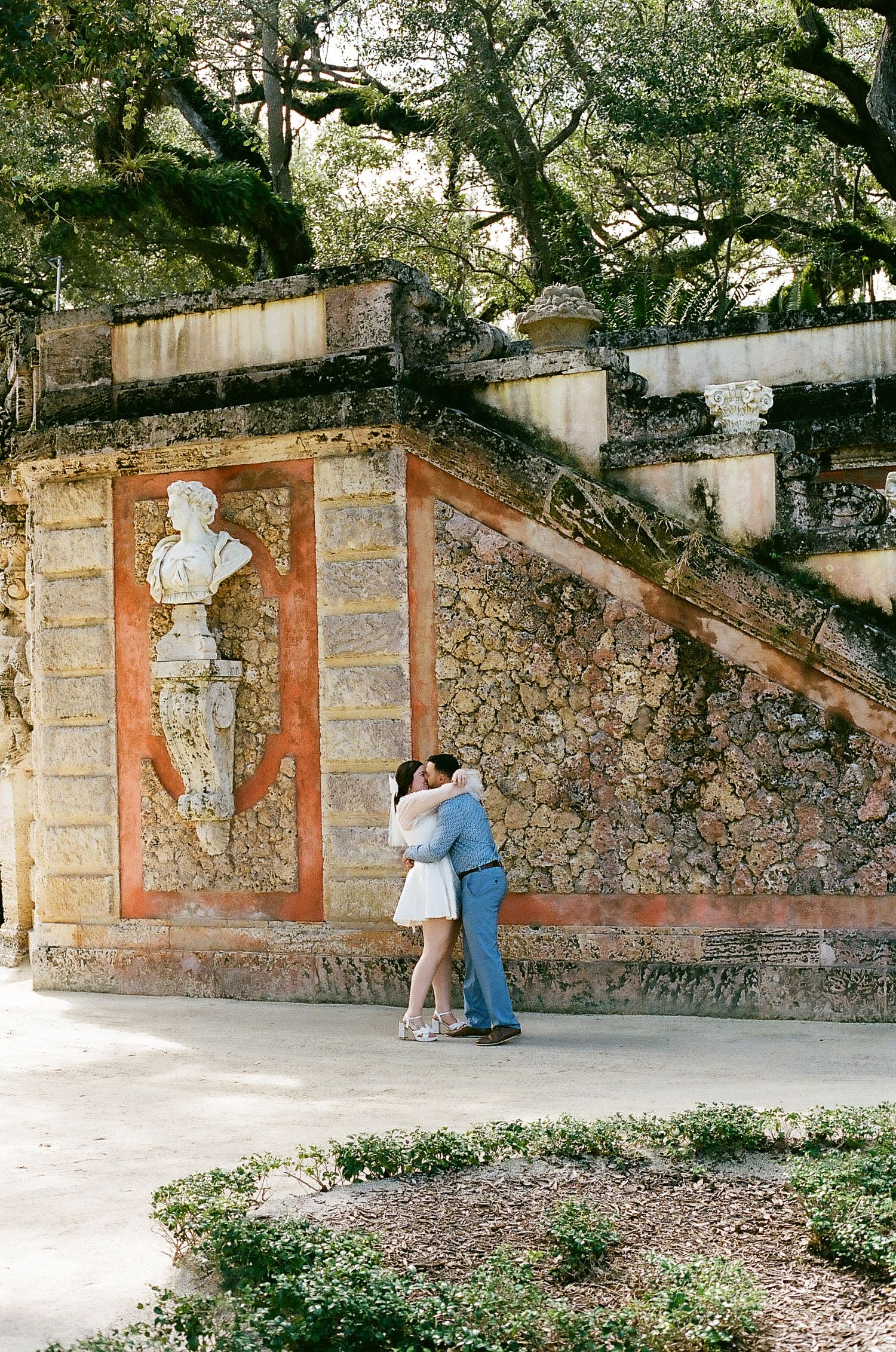 A romantic couple shares a tender moment against a stone wall with decorative niches under a canopy of oak trees.