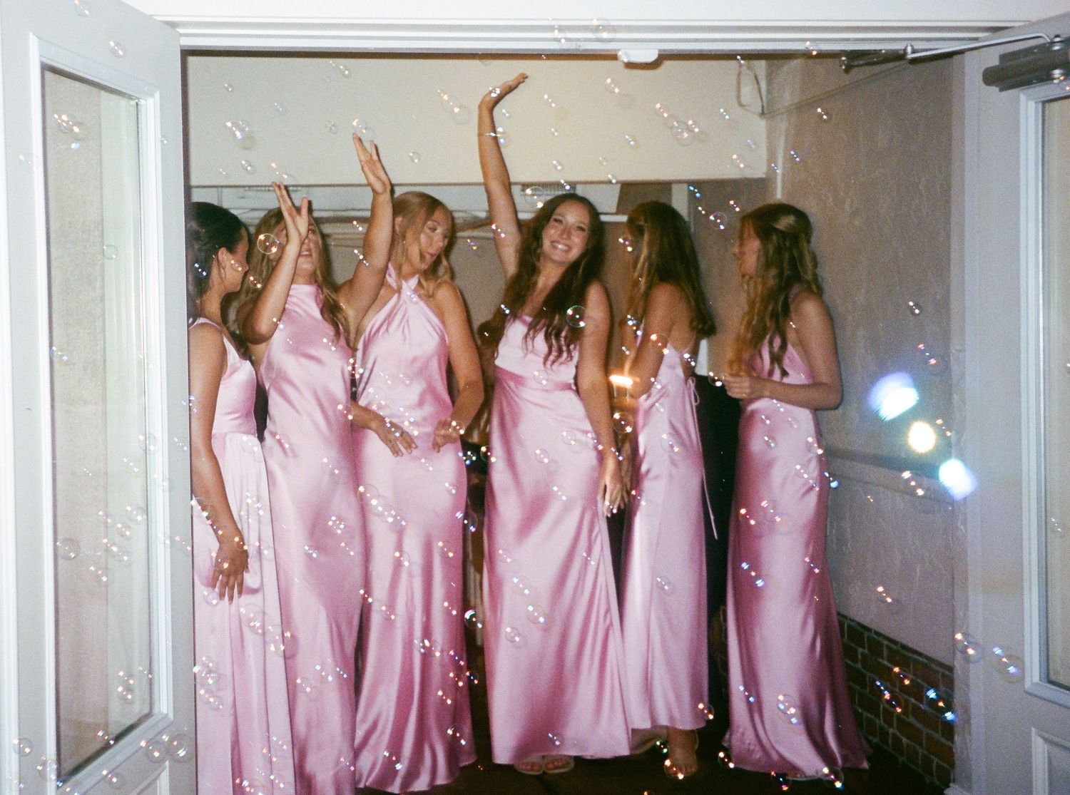 A group of bridesmaids in matching pink dresses pose together in a wedding venue hallway.