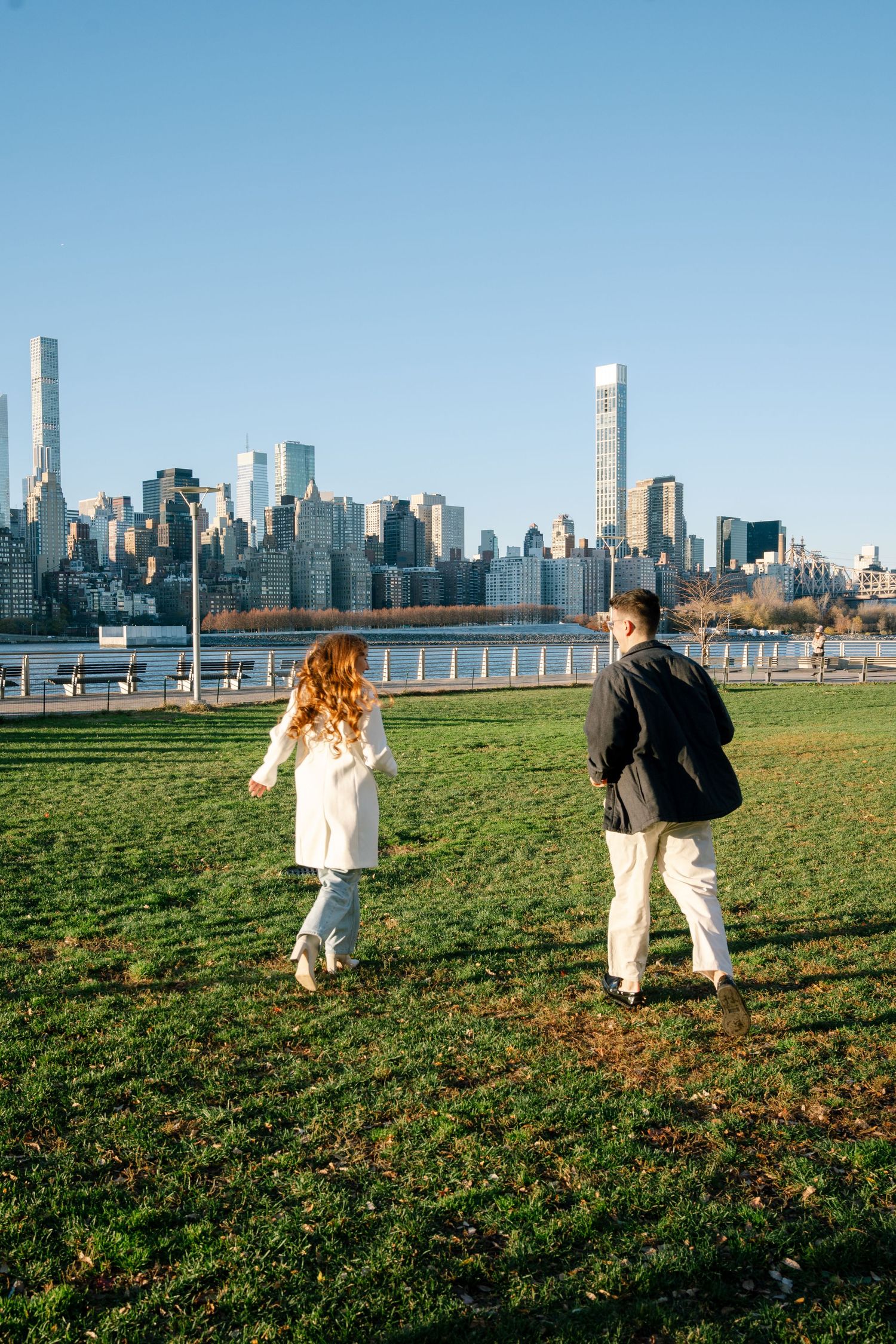 Two people walk and play on a grassy field with the New York City skyline visible in the background on a sunny day.