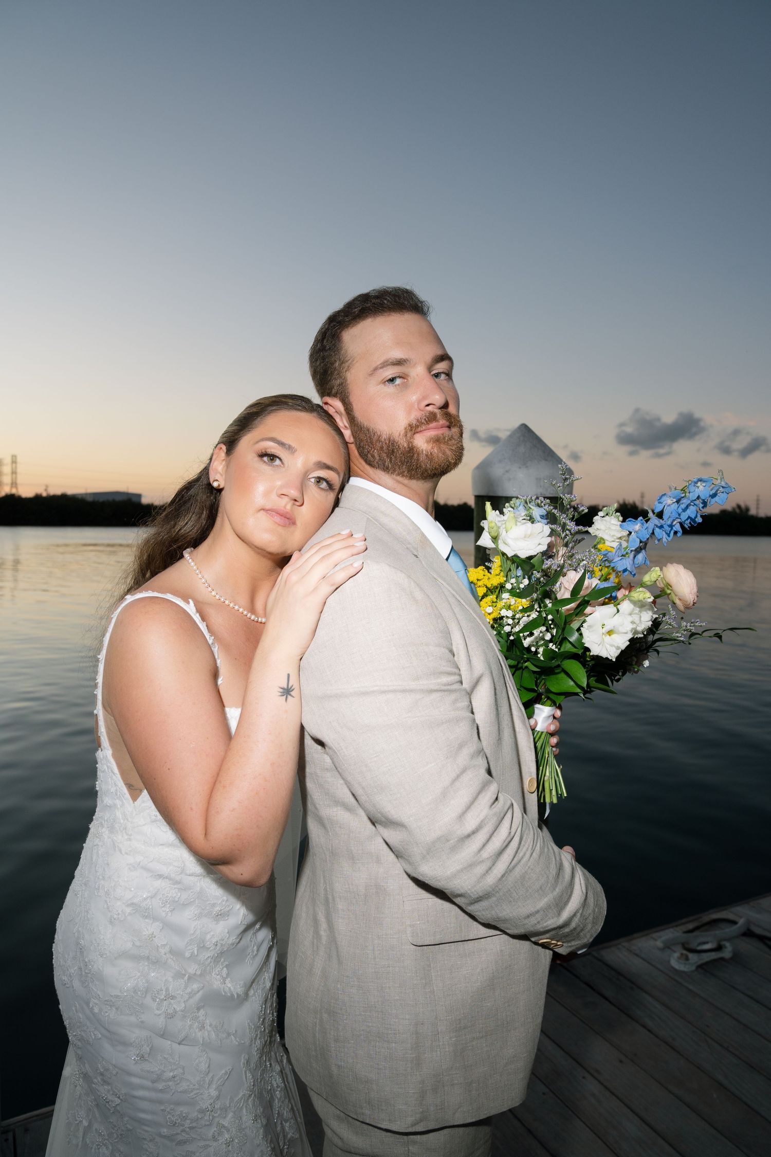 Newlywed couple posing on wooden dock at sunset with water and sky in background.