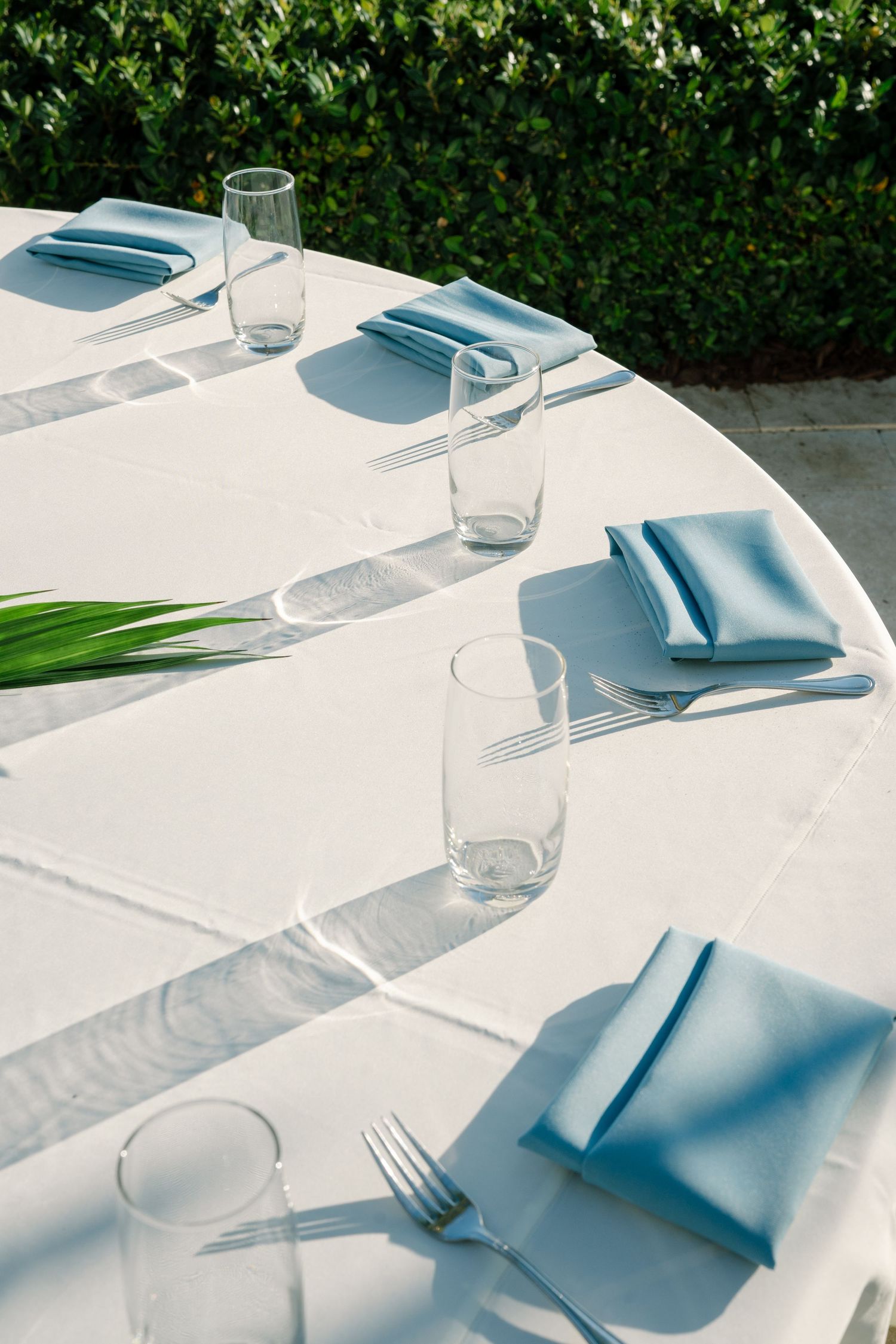 Clean white table settings with light blue napkins and glassware arranged on a bright outdoor dining table.