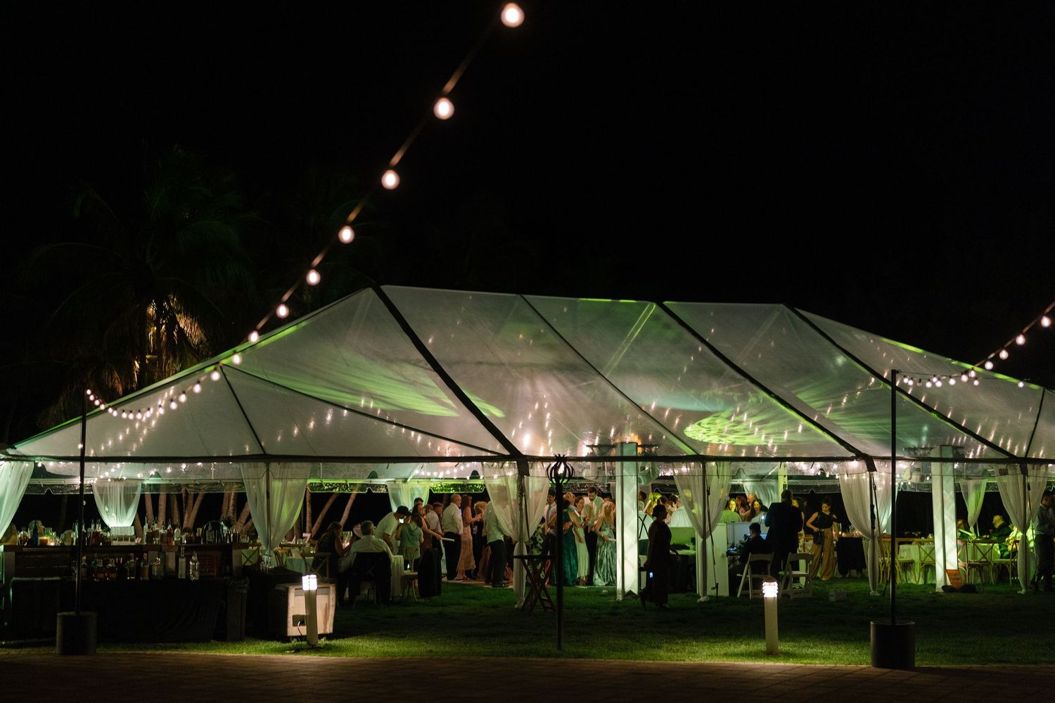 Elegant clear wedding reception tent glowing at night with string lights and guests celebrating inside.