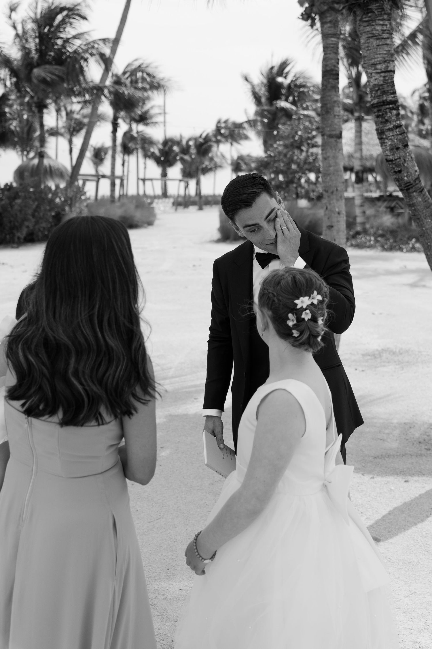 Emotional black and white photo of groom wiping tears during first look with bride on beach.