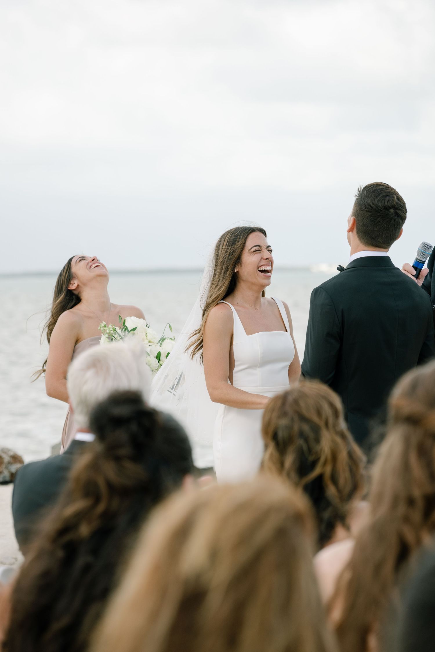 Laughing bride standing at outdoor waterfront ceremony with bridesmaid and officiant on an overcast day.