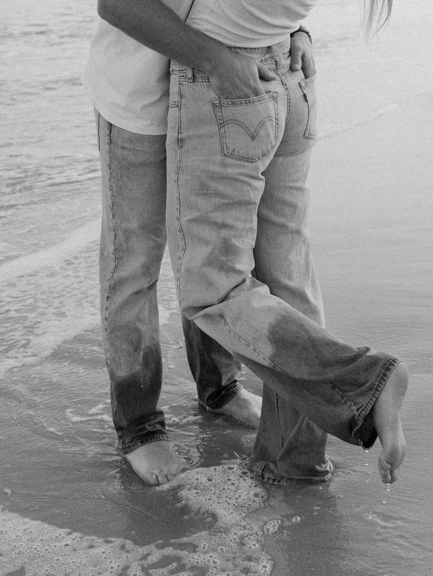 A series of black and white photographs showing legs in jeans standing in shallow ocean waves at the beach.