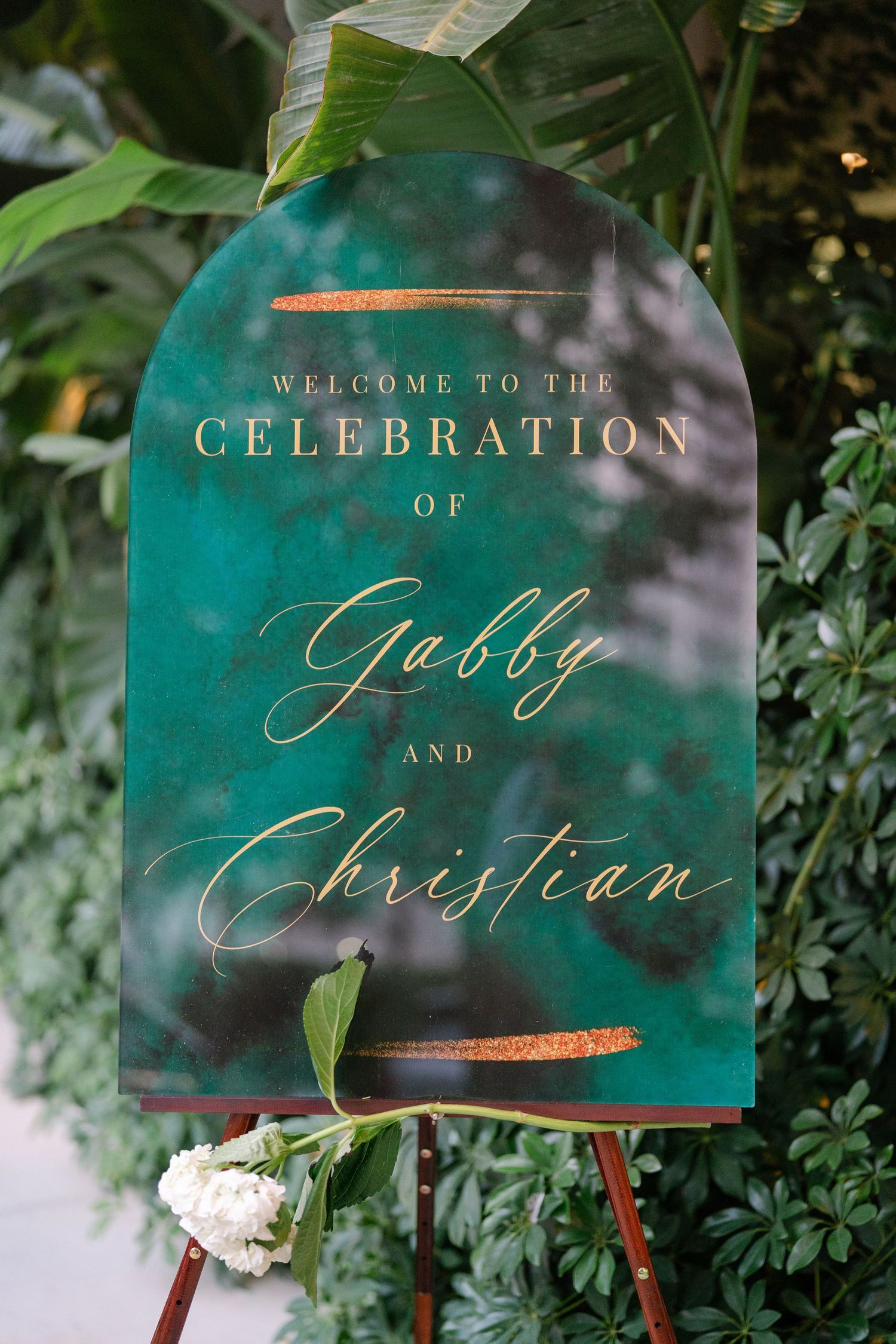 An elegant emerald green wedding welcome sign with gold lettering displayed on an easel among foliage.