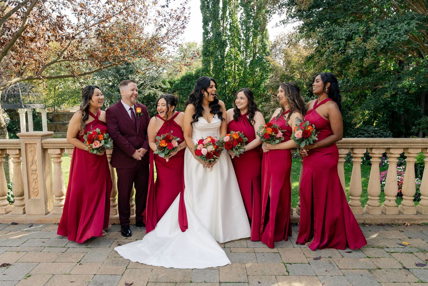Wedding party in burgundy dresses and formal attire poses together on stone steps outdoors.