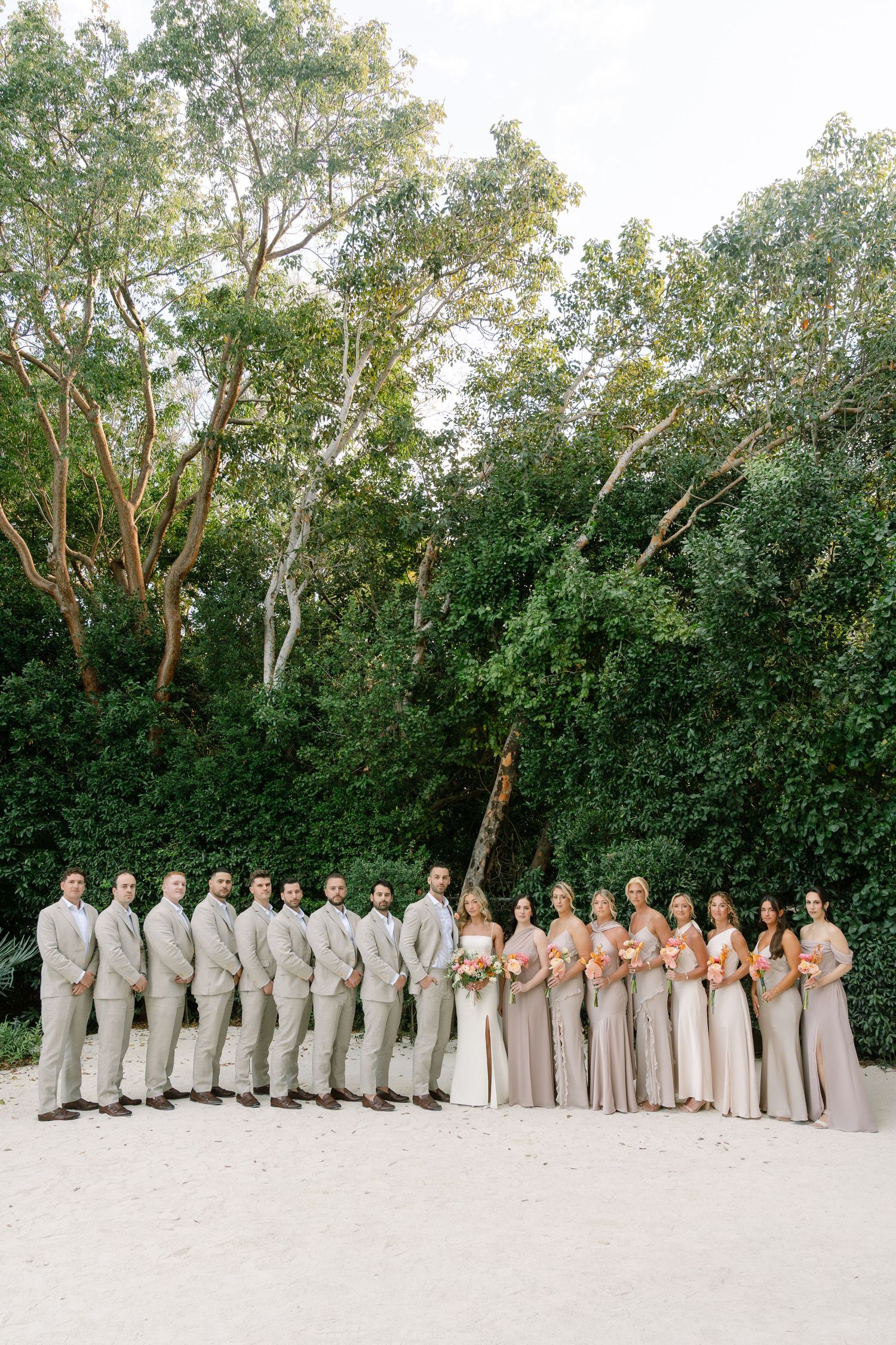 Wedding party in neutral tones lined up for formal photo against tropical greenery backdrop.