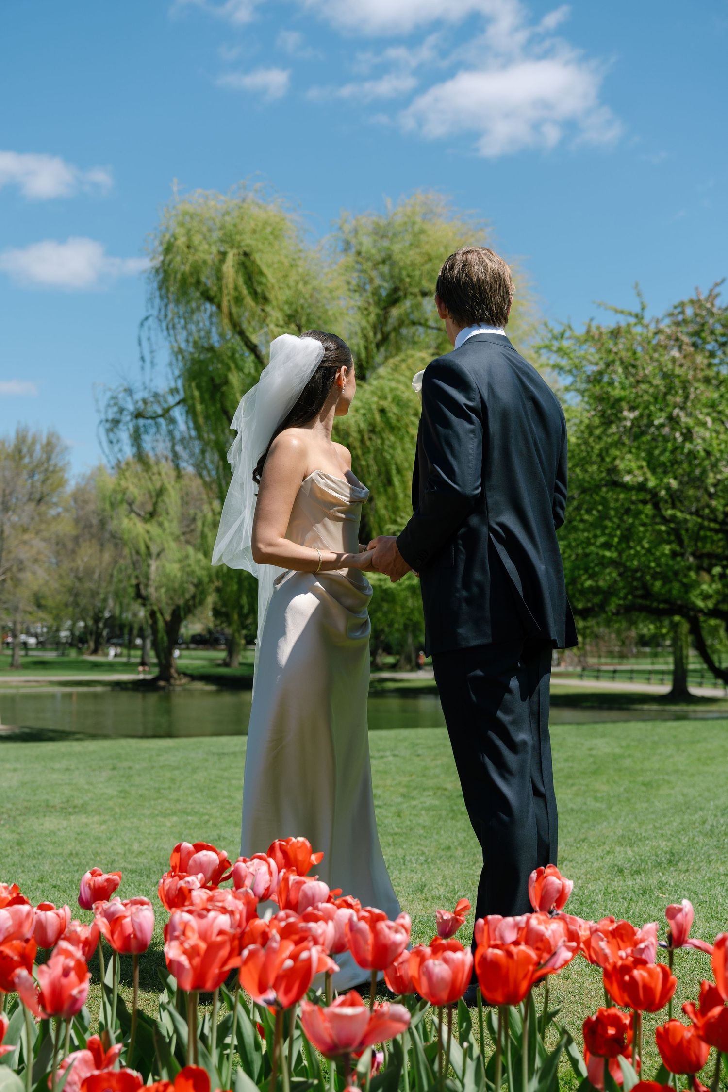 Bride and groom holding hands in a lush green park with red tulips in the foreground on a sunny wedding day.