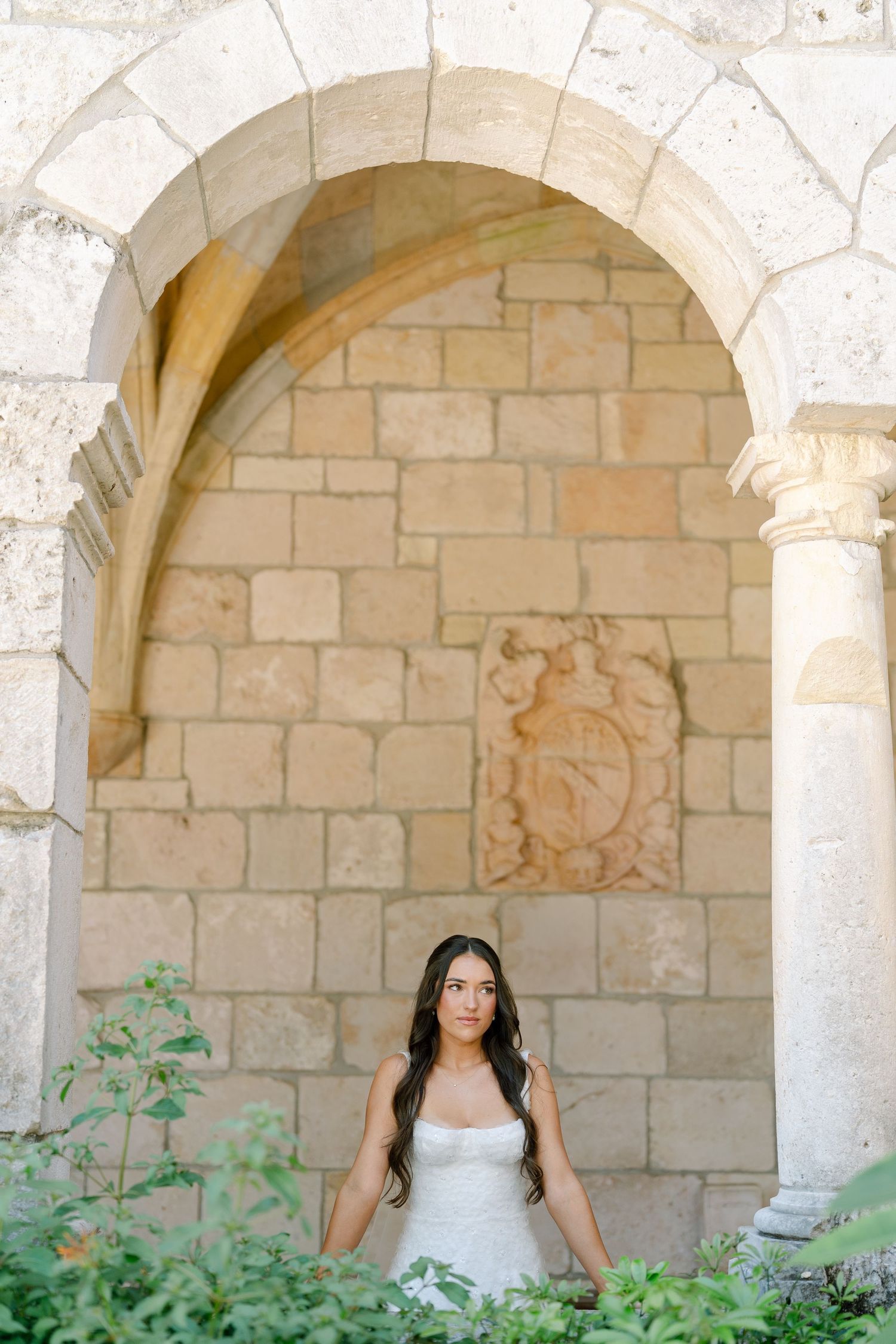 Bride in strapless white gown poses under ornate stone arch with carved relief in historic venue.