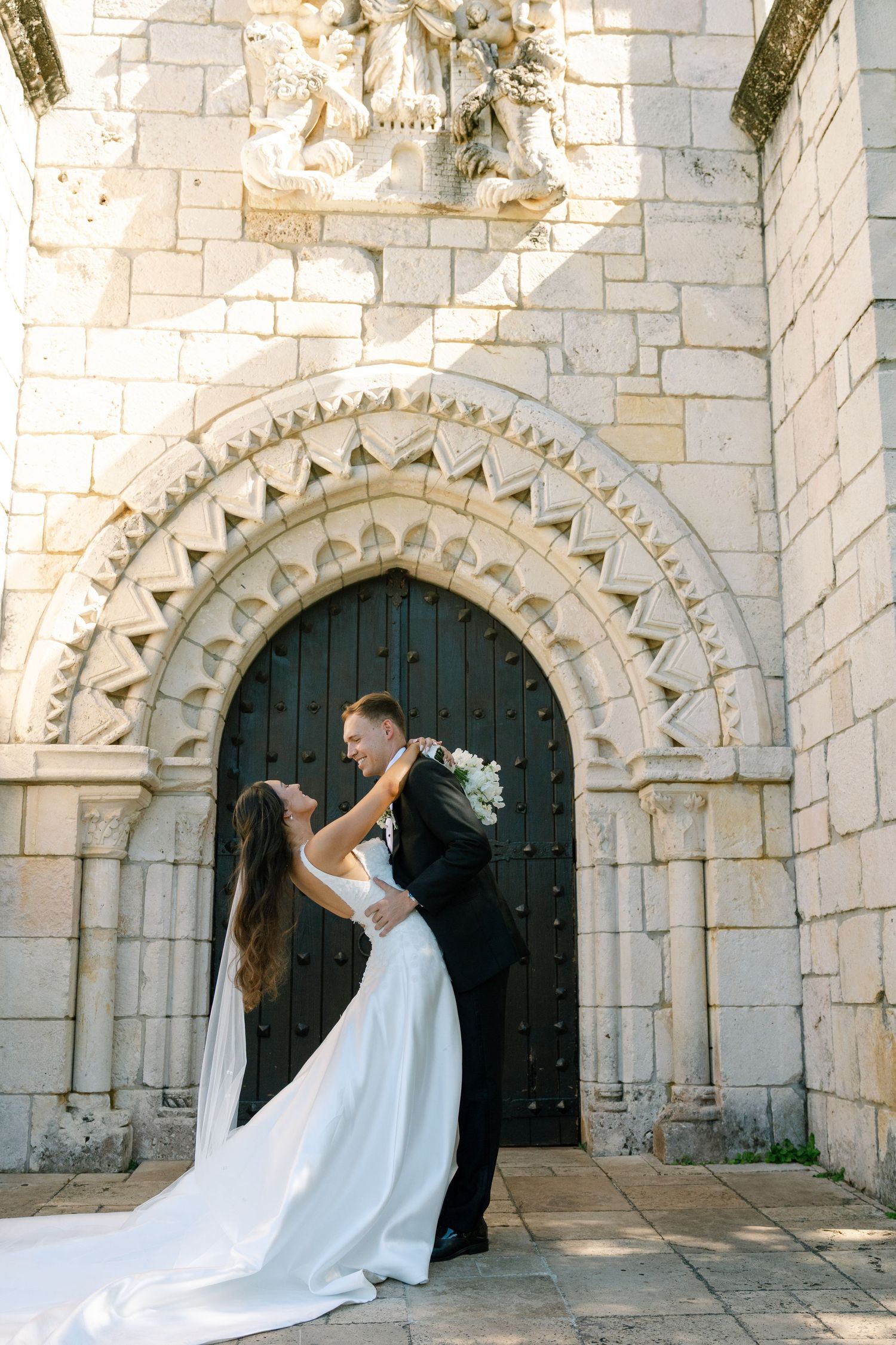 Bride and groom dancing romantically before ornate arched stone church doorway in sunlight.