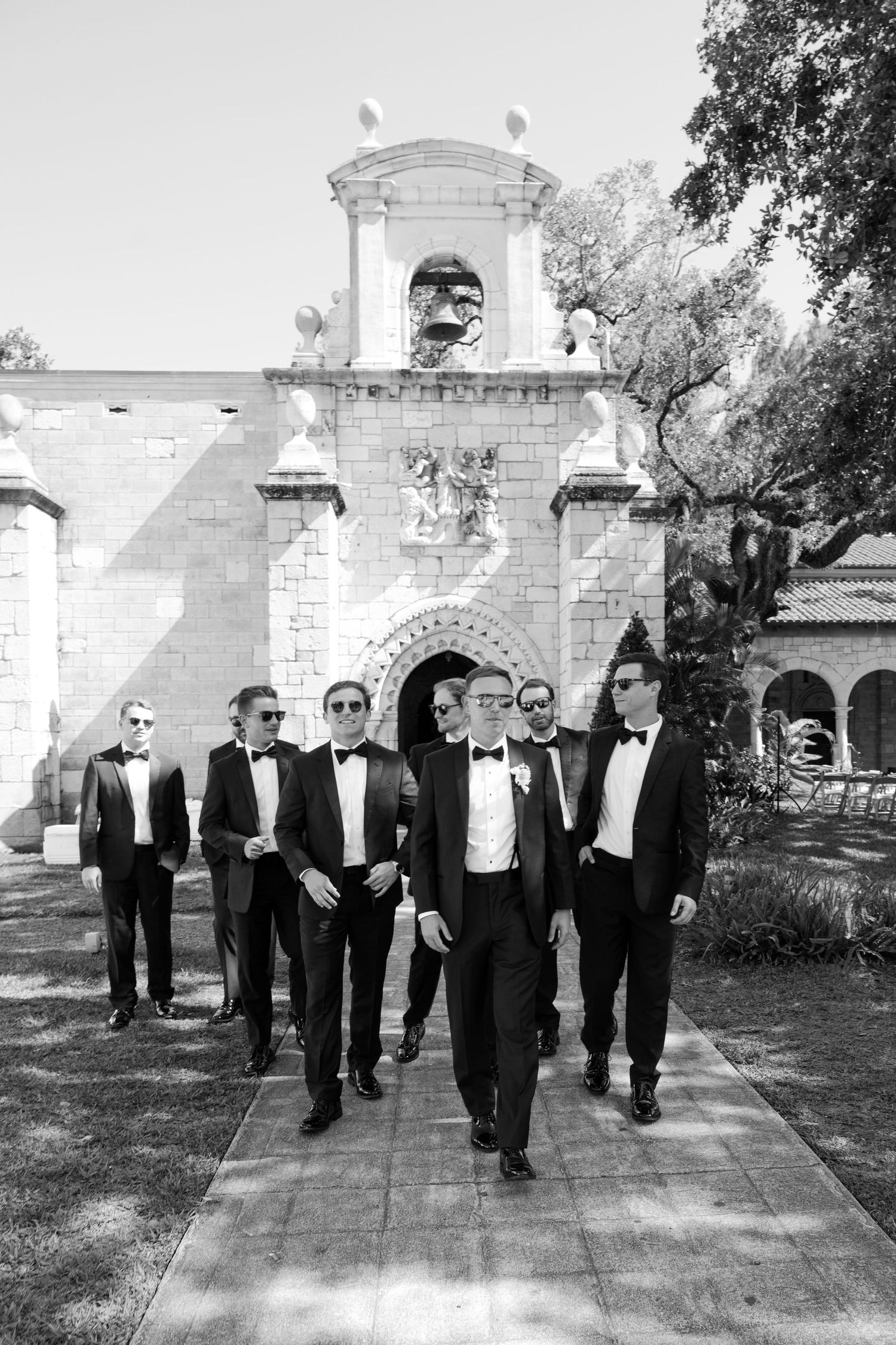 Black and white photo of groom leading groomsmen in tuxedos walking from historic stone chapel entrance.