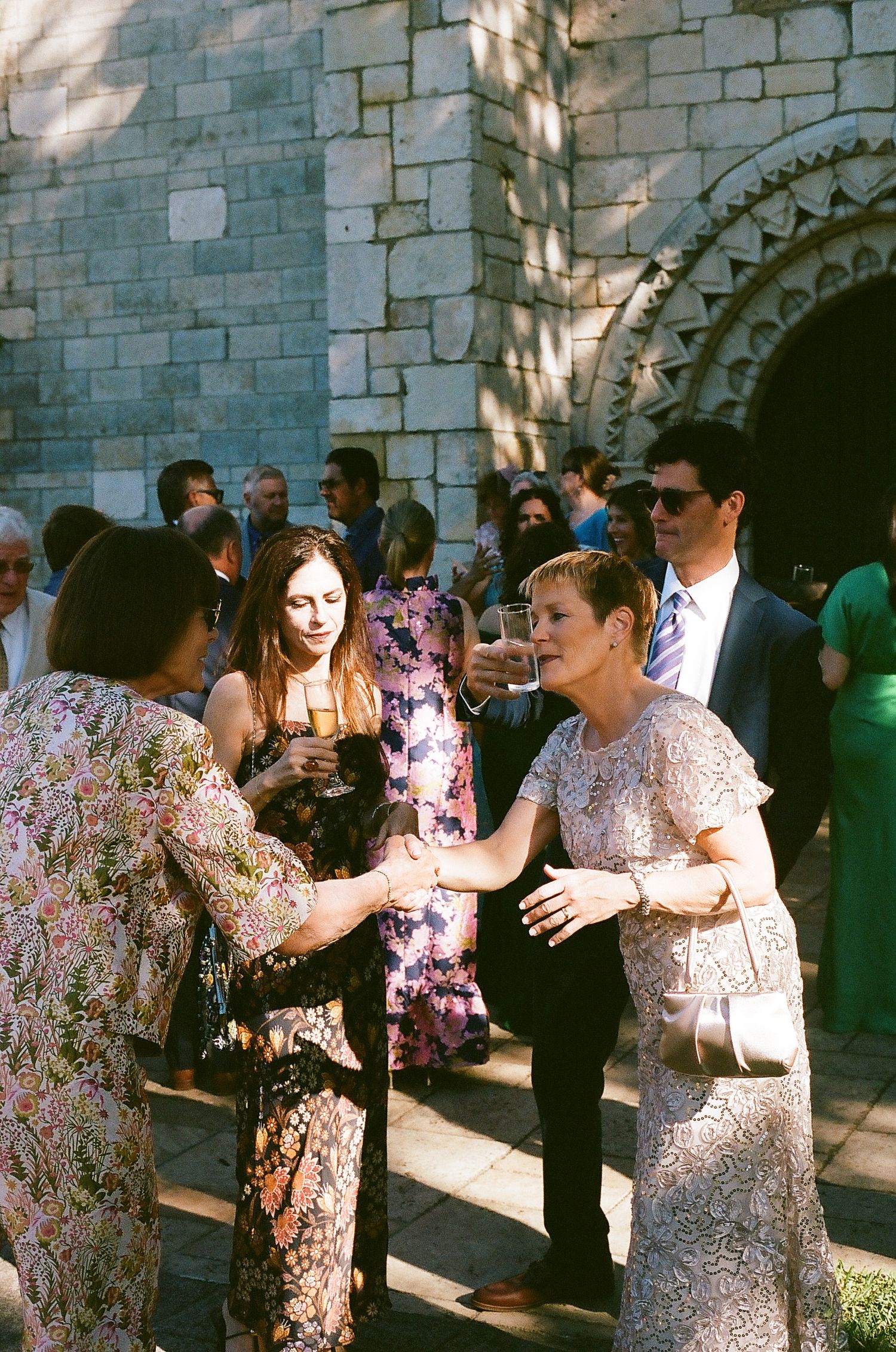 Elegantly dressed wedding guests mingle and shake hands outside a historic stone church during a sunny outdoor reception.