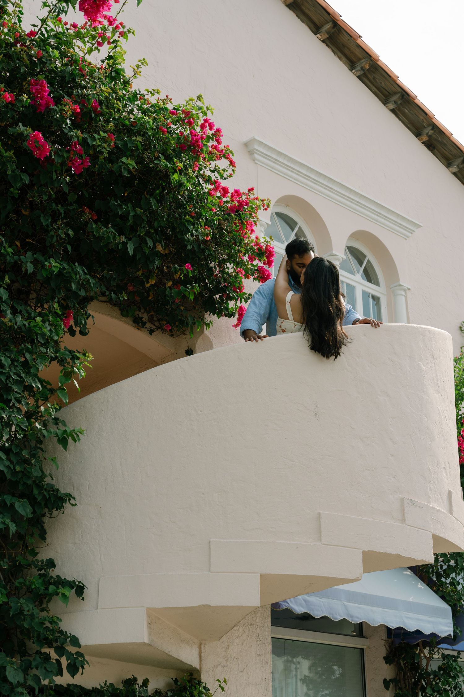 Couple sharing a romantic kiss on a white curved balcony adorned with vibrant pink bougainvillea flowers.