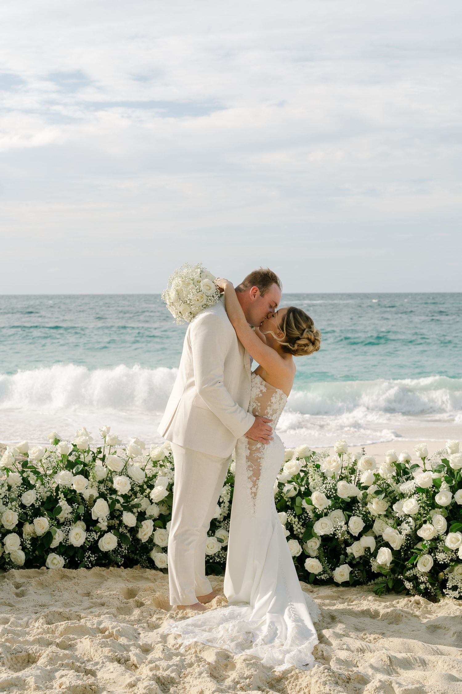 Couple sharing first kiss at beach wedding ceremony with white floral backdrop and ocean waves.