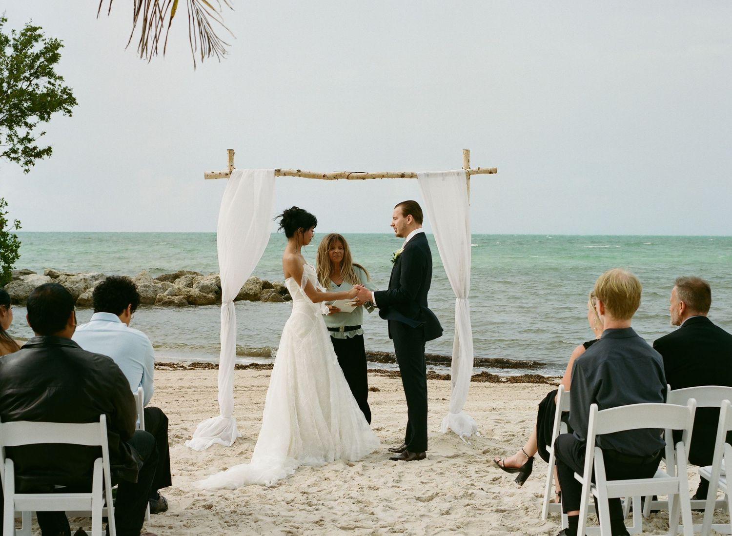 Couple exchanges vows at a romantic outdoor beach wedding ceremony under a draped wooden arch by the ocean.
