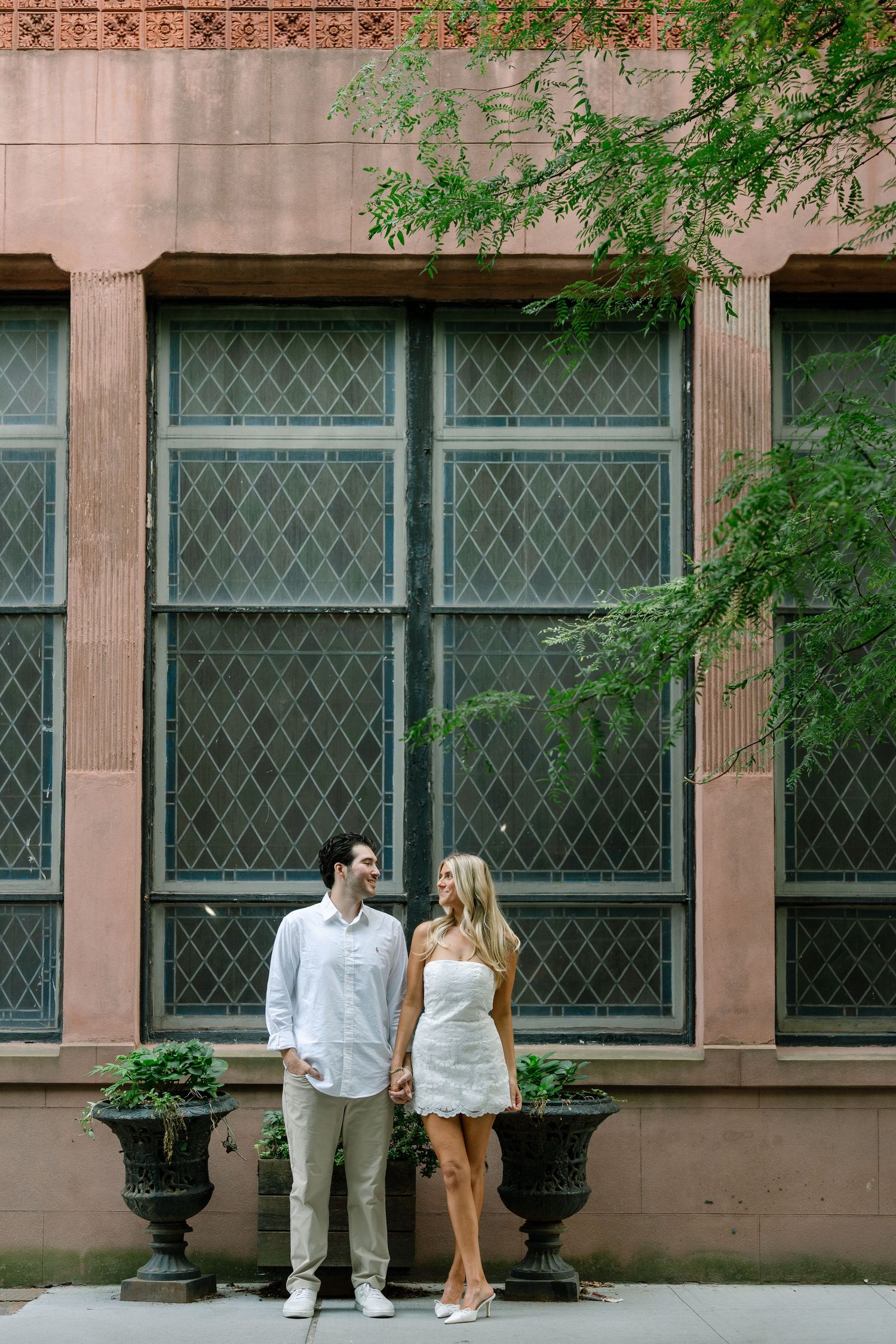 Couple holding hands in white outfits standing before a large latticed window on a brick building with decorative planters.