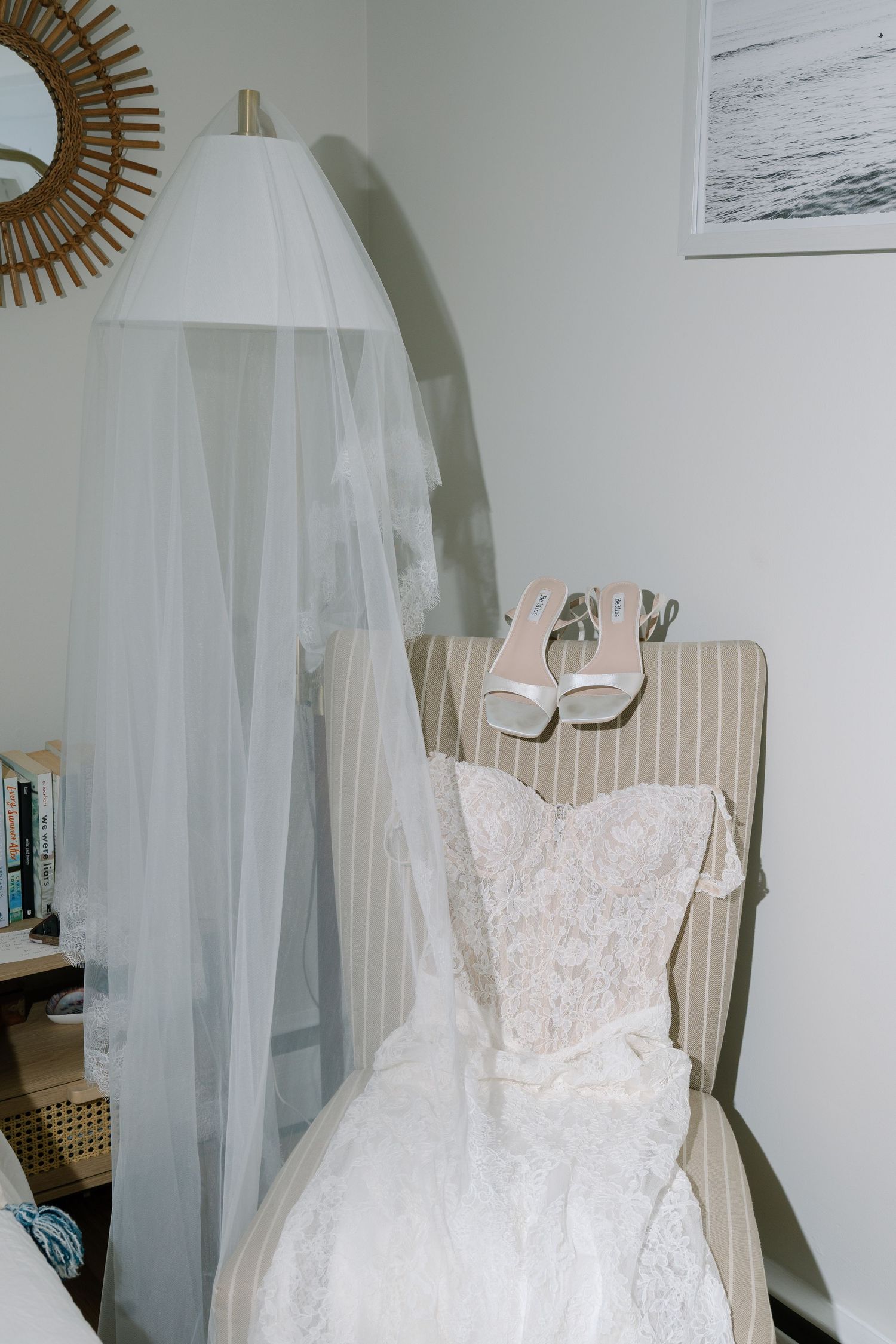White lace wedding dress and veil displayed on striped chair with bridal heels in elegant bedroom setting.