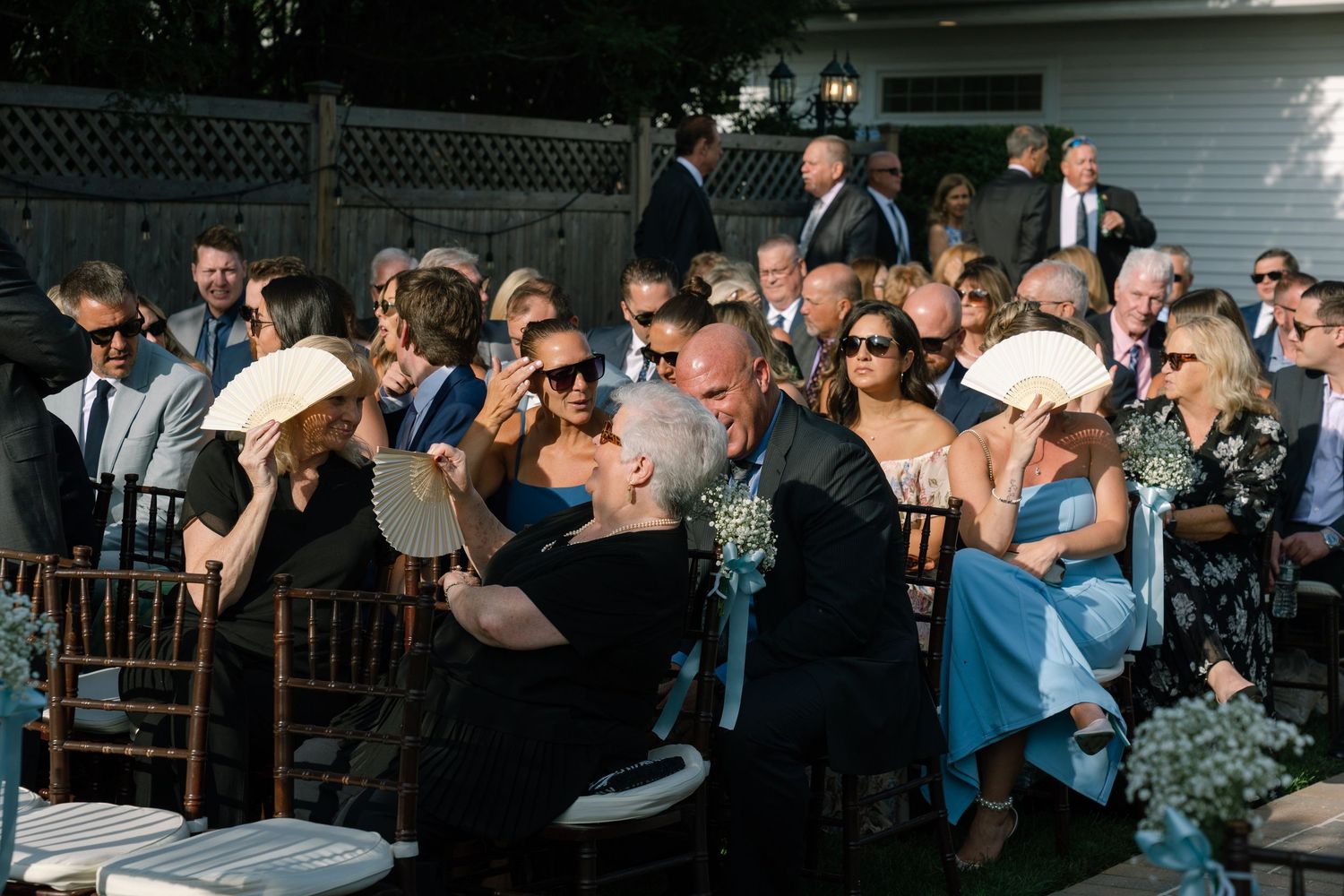 Outdoor wedding guests seated in chairs, socializing and smiling during a sunny ceremony reception event.