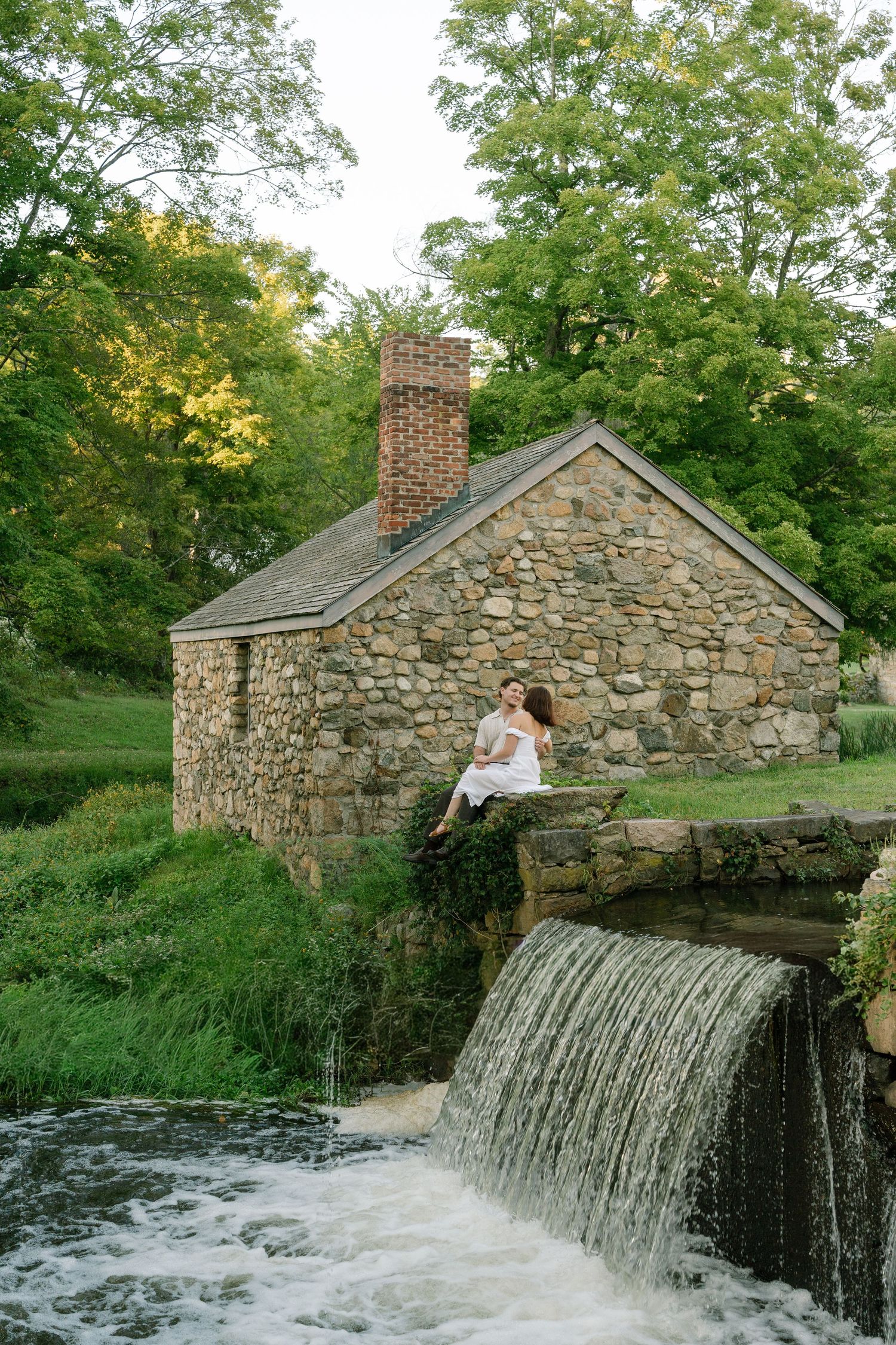 Couple sitting on stone wall beside rustic mill waterfall surrounded by lush green trees.