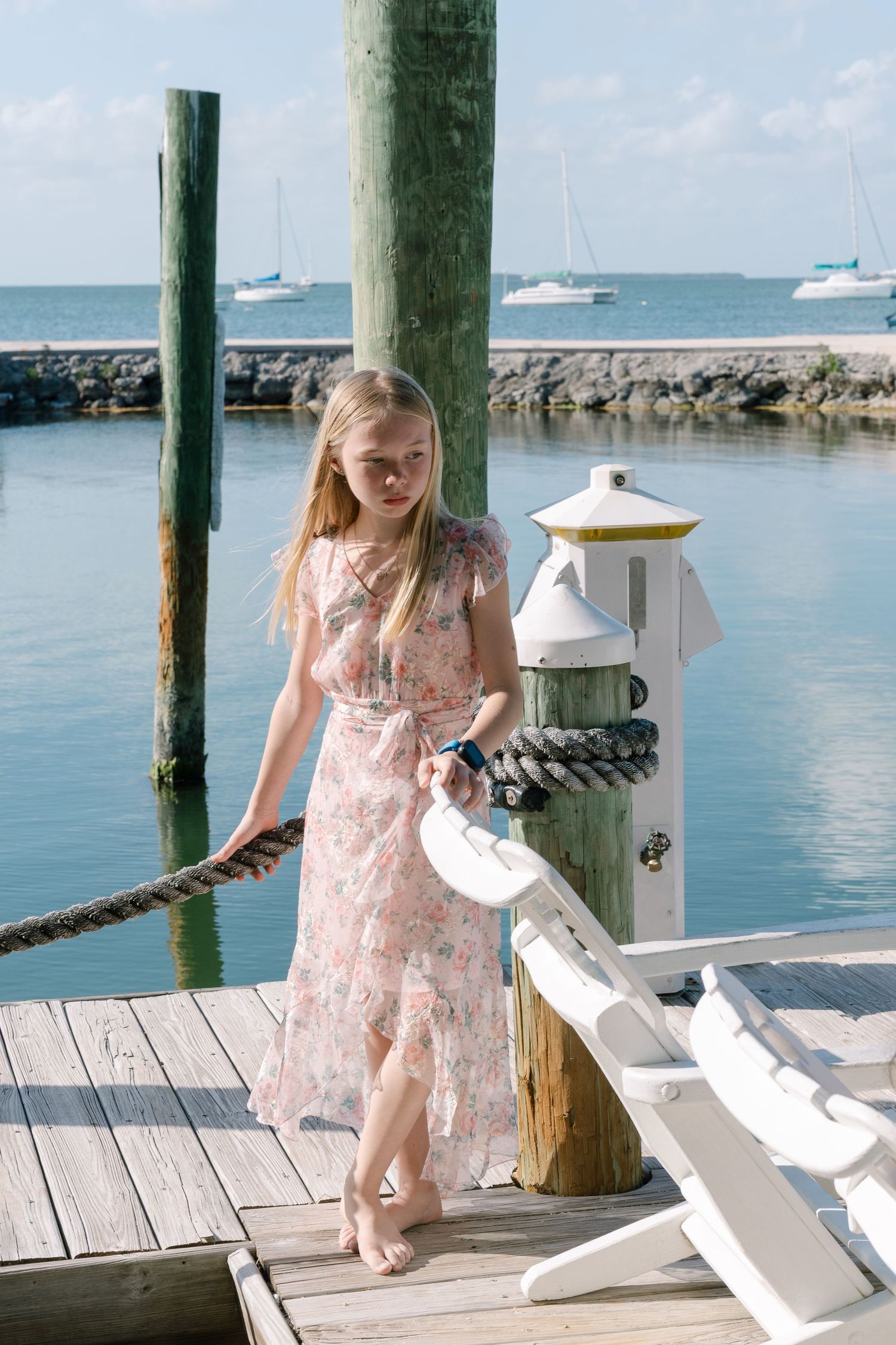 A young blonde girl in a floral pink dress stands barefoot on a wooden dock with sailboats in the background.