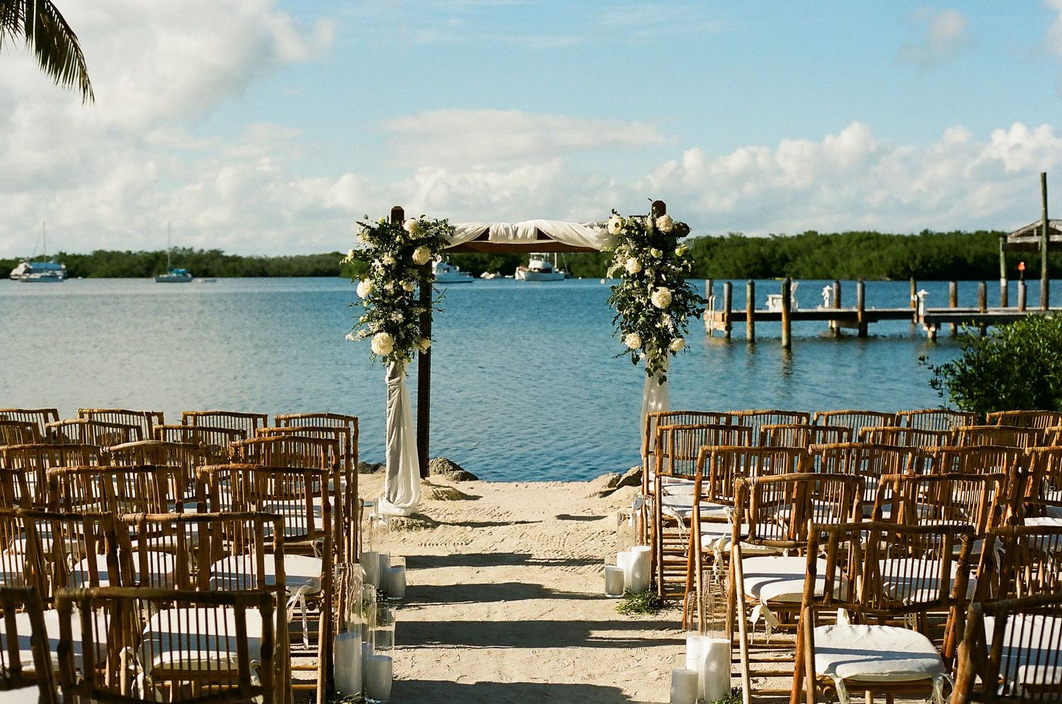 Wooden chairs arranged on a beach for a tropical waterfront wedding ceremony with palm trees and ocean views.