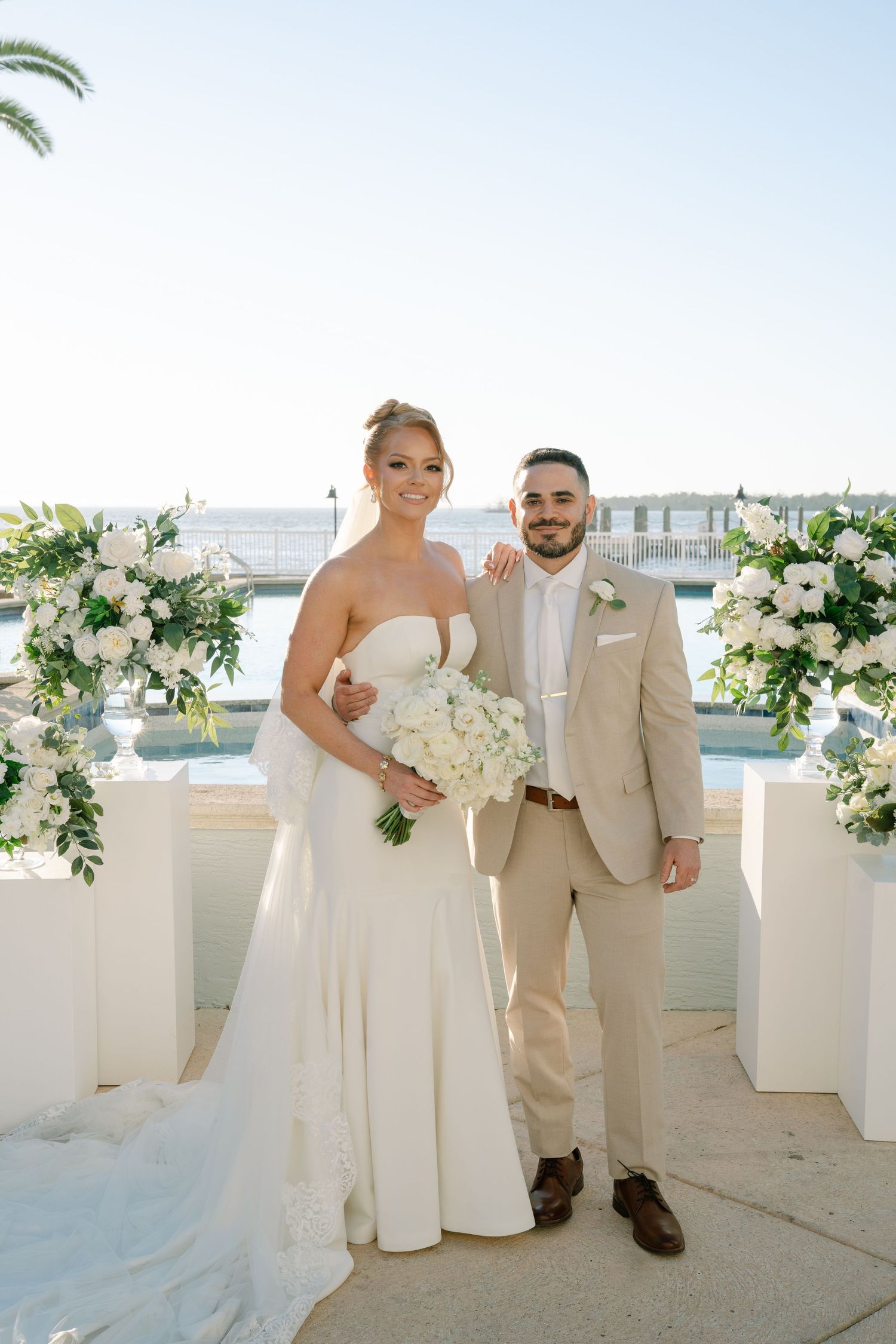 A wedding couple poses on a waterfront terrace decorated with white floral arrangements on a sunny day.