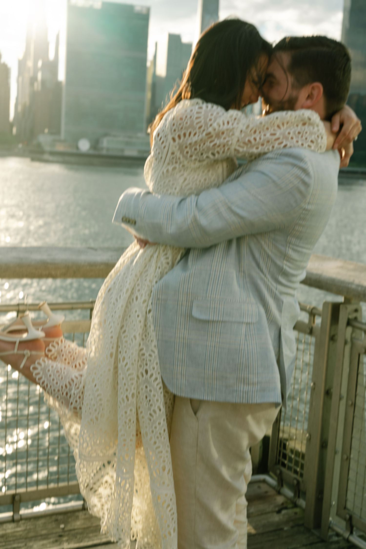 Couple embracing and kissing on a waterfront deck with a city skyline at sunset, dressed in elegant cream outfits.