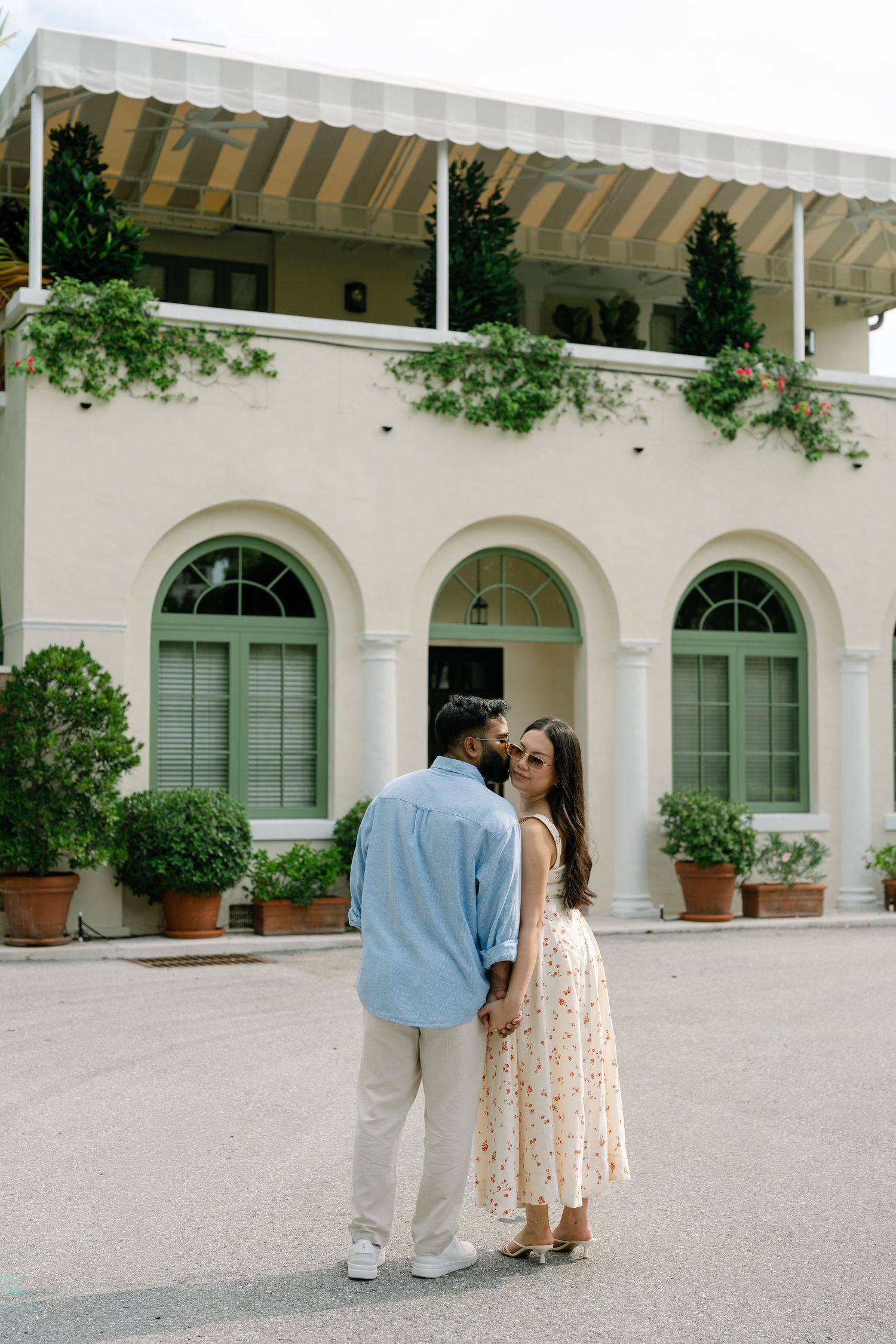 Couple holding hands and sharing a kiss in front of an elegant white Mediterranean-style building with arched windows and lush greenery.