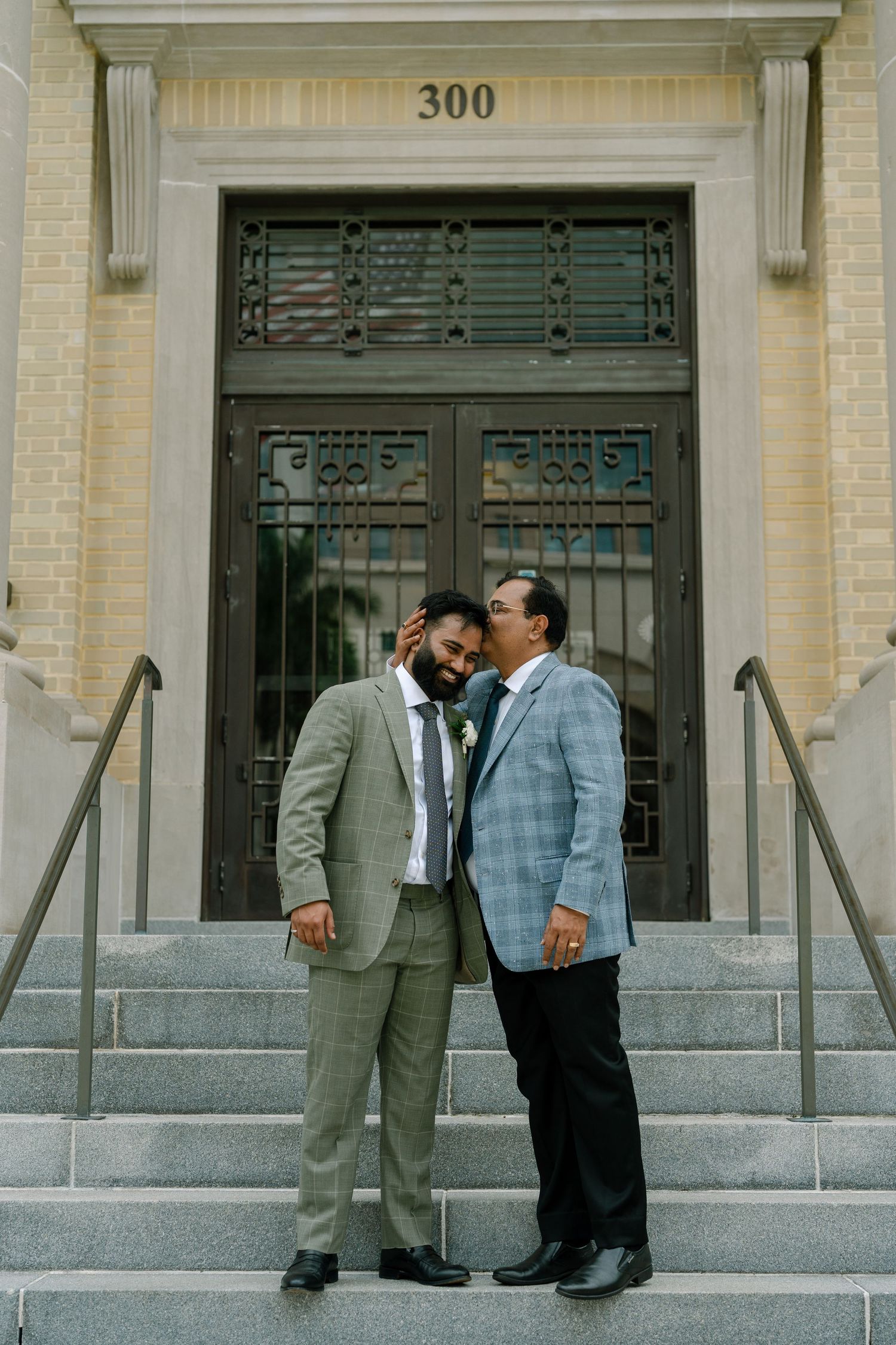 Two grooms share a tender moment on steps outside building 300, one in gray suit, other in plaid blazer.
