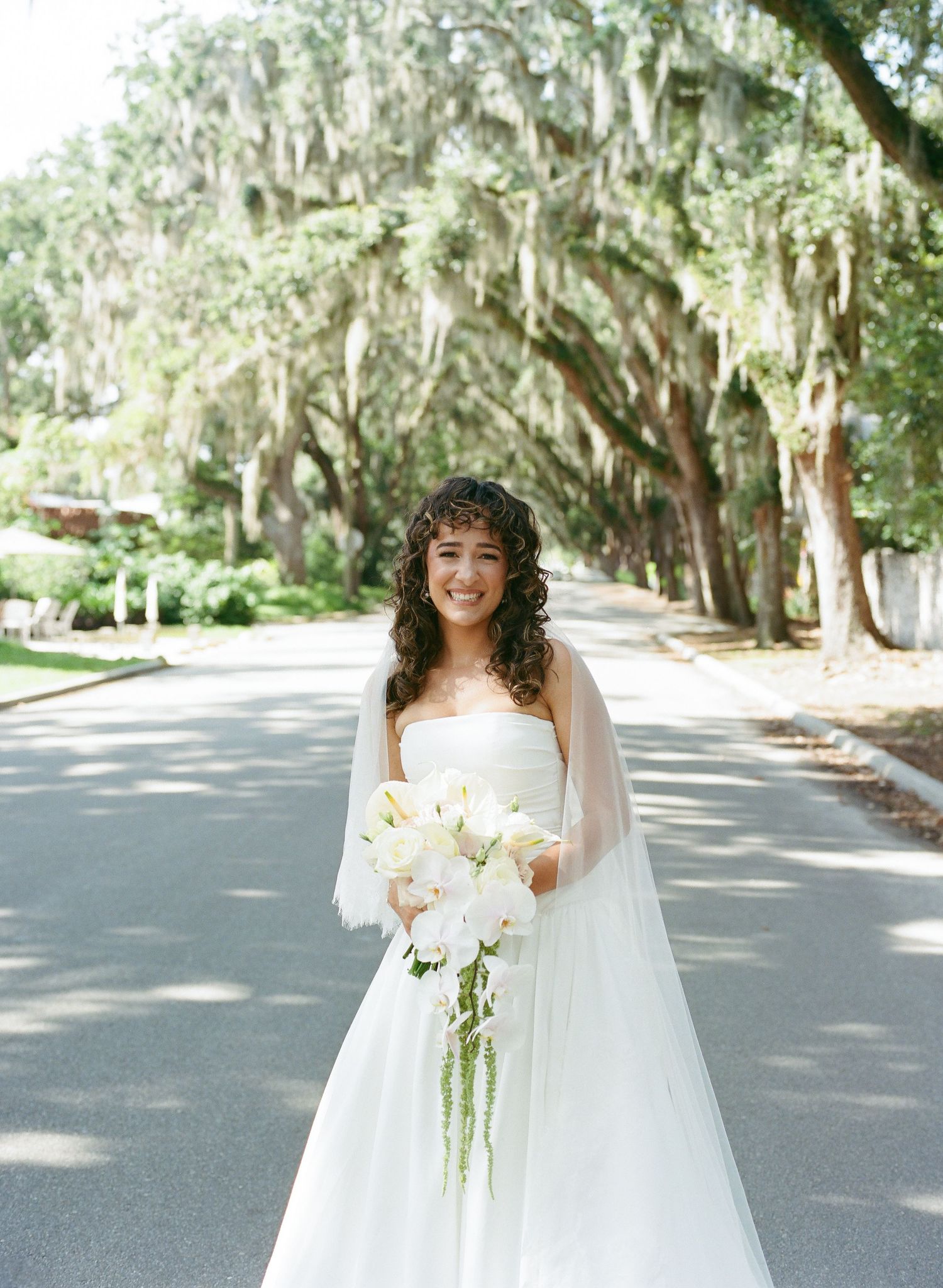 Bride in flowing white gown poses joyfully on a tree-lined path with Spanish moss overhead.