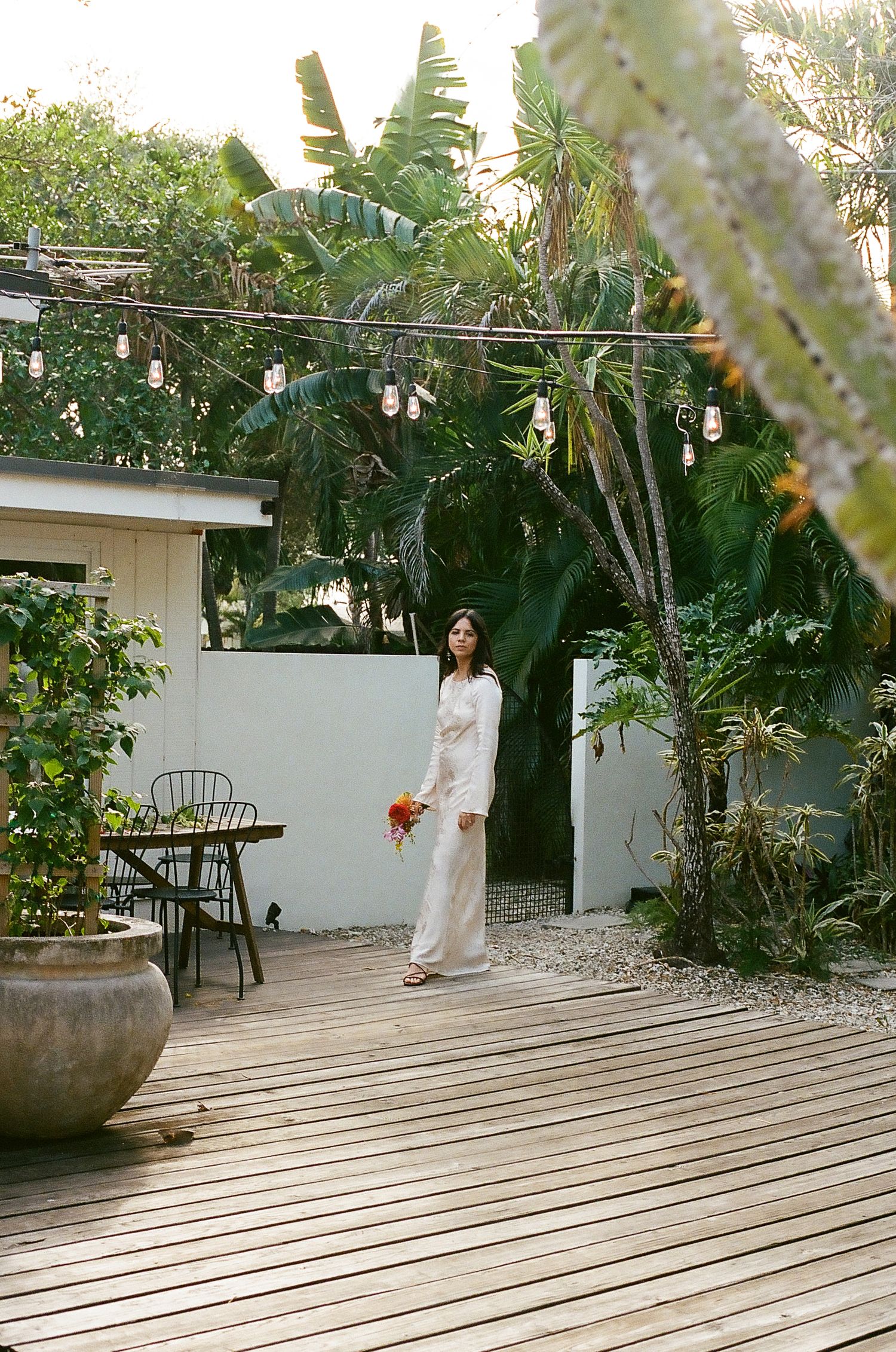 A person in a white dress stands on a wooden deck surrounded by lush tropical plants and garden furniture.
