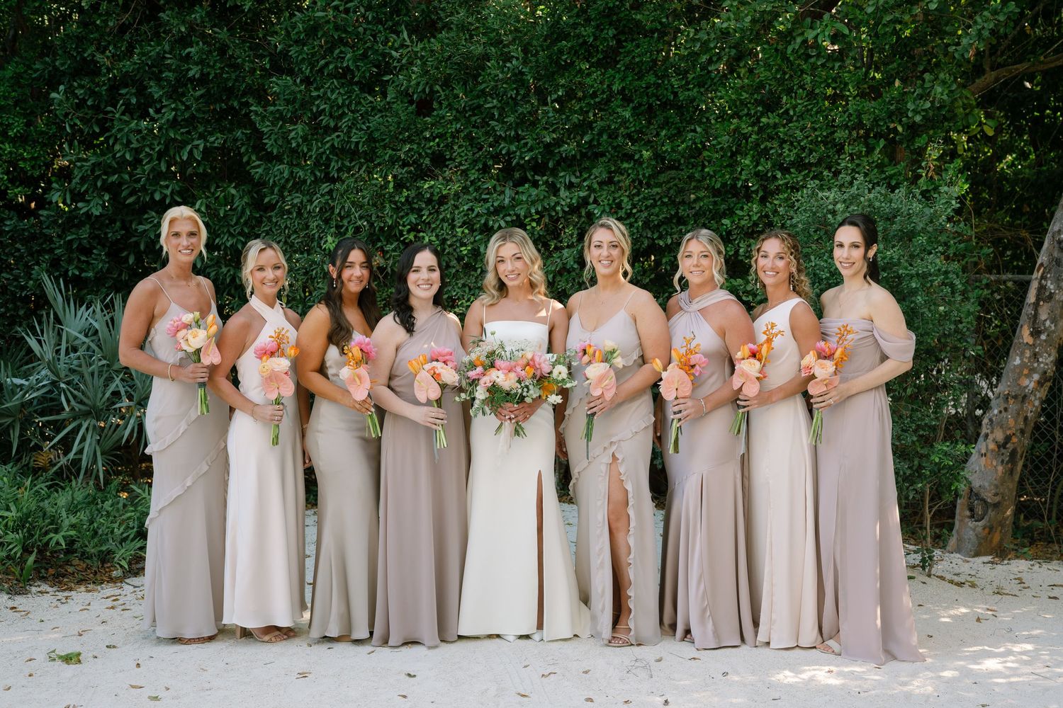 Wedding party in neutral-toned dresses posing together against lush green foliage backdrop.