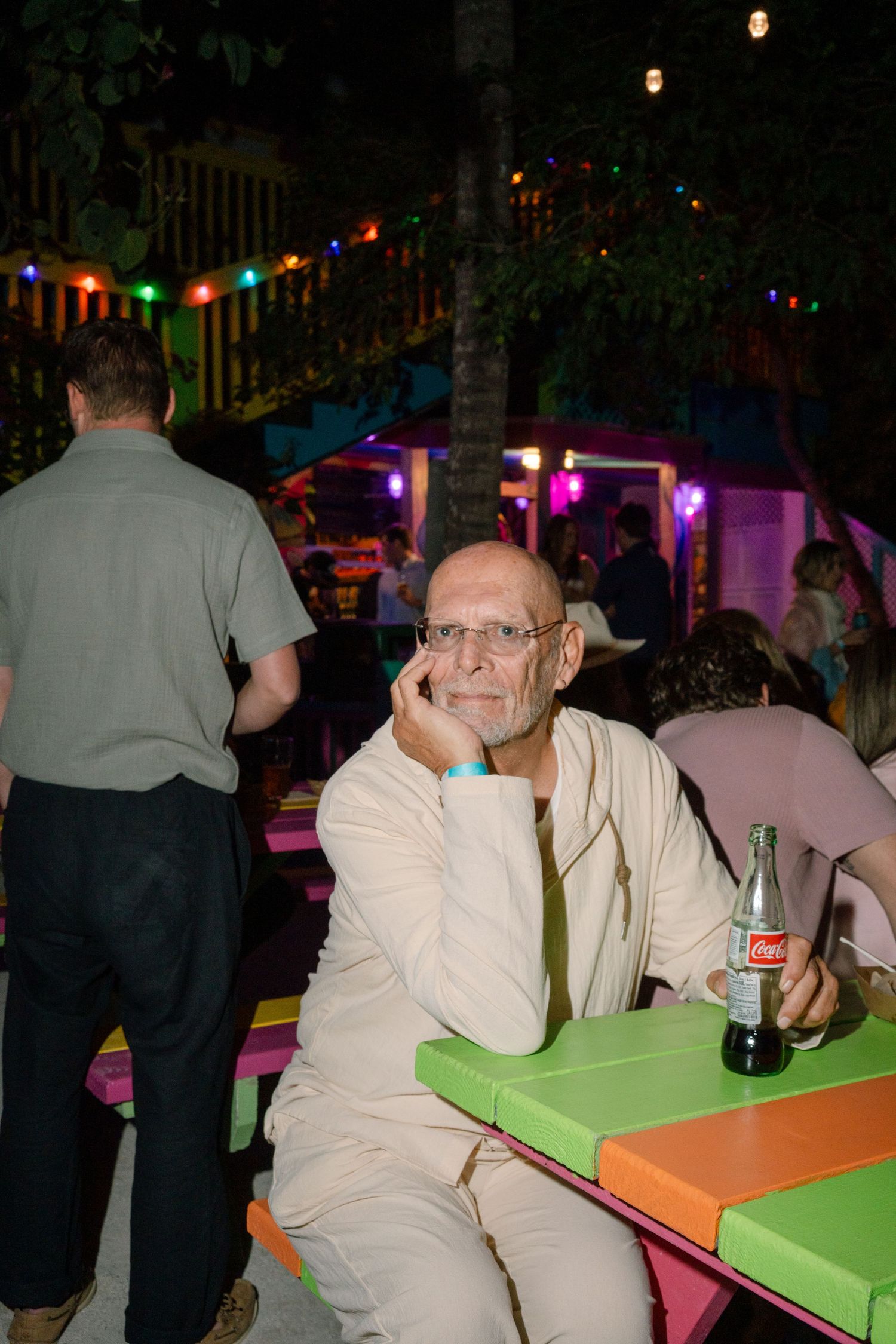 Person in light colored clothing sitting at colorful outdoor table with festive lighting.